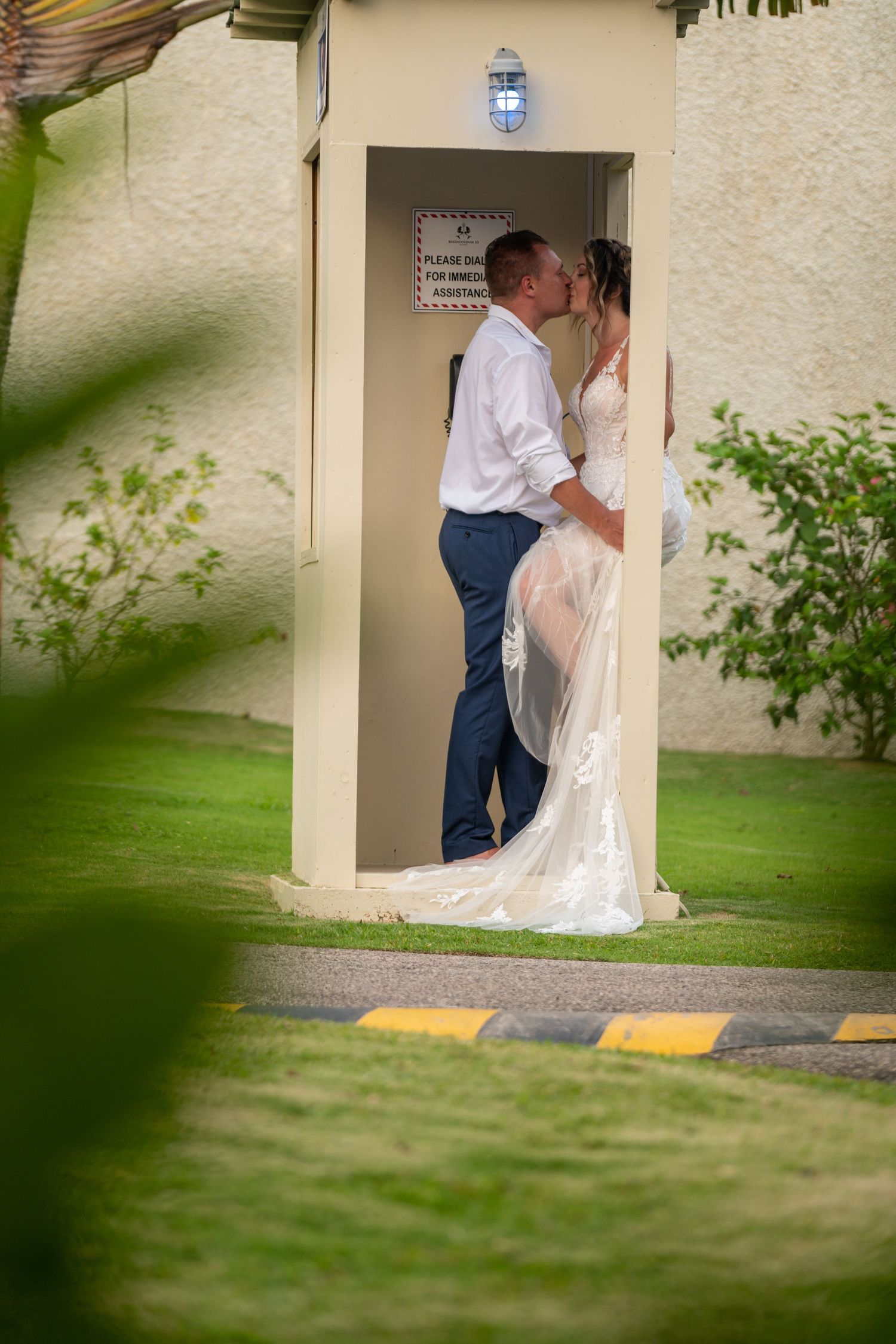 A couple shares an intimate moment against a cream-colored telephone booth with green foliage in the foreground.