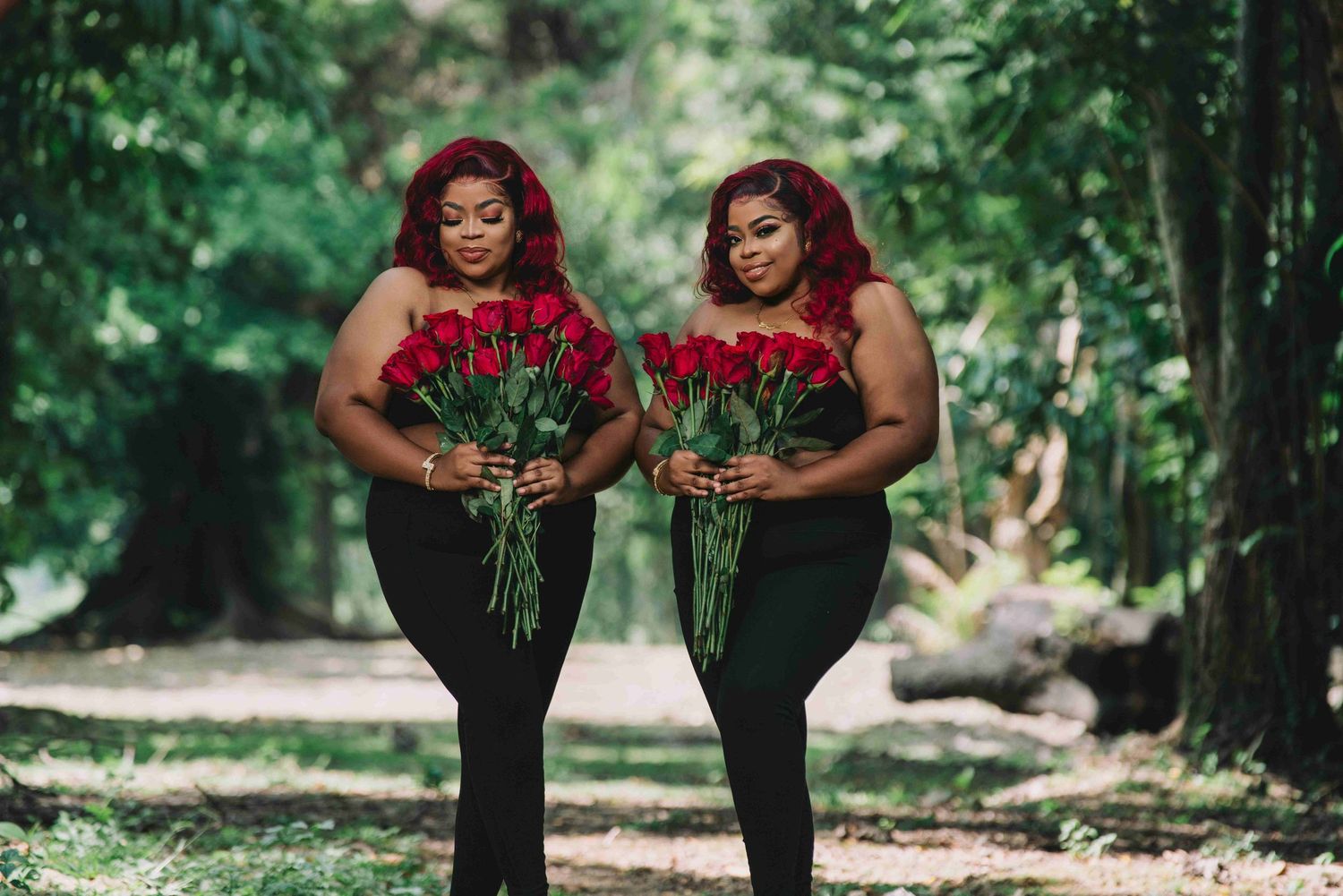 Two women in black outfits holding red roses pose together in a lush green forest setting.