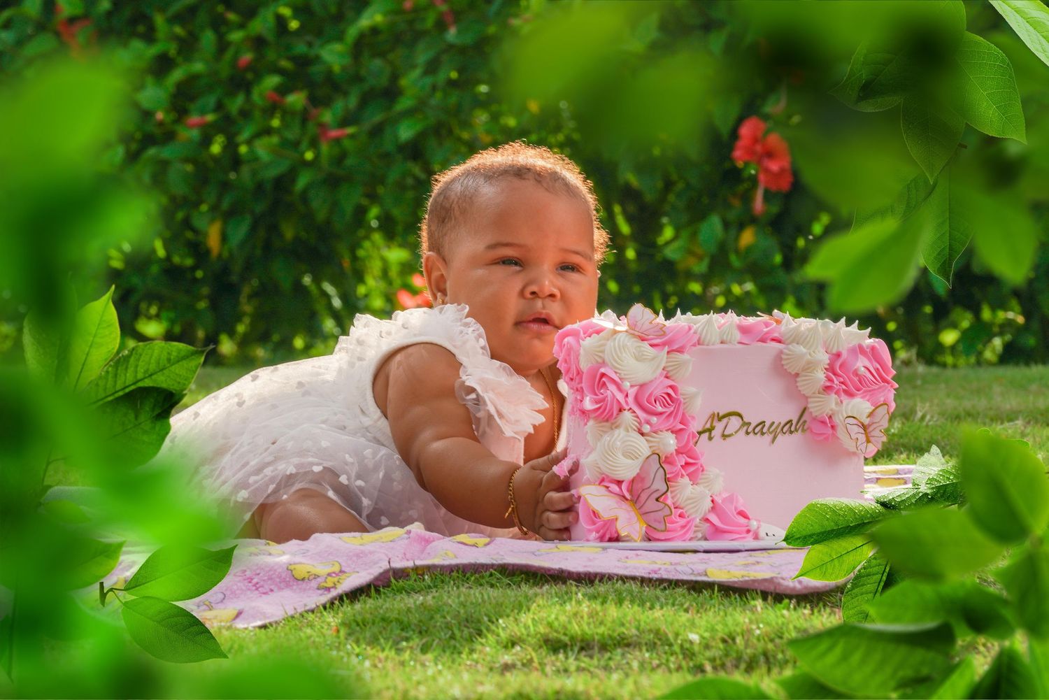 Infant in a pink dress lying on a blanket outdoors surrounded by lush green foliage and smiling joyfully.