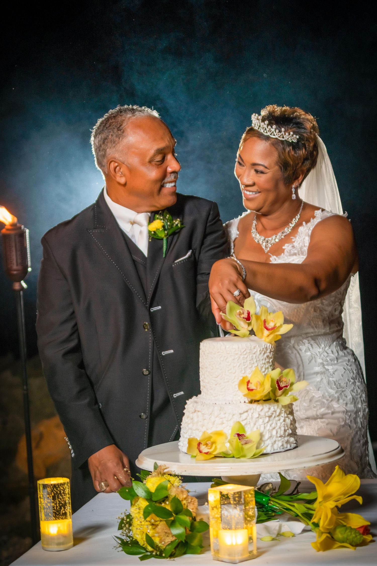 Wedding couple smiling and laughing while cutting their white wedding cake decorated with yellow calla lilies and candles.