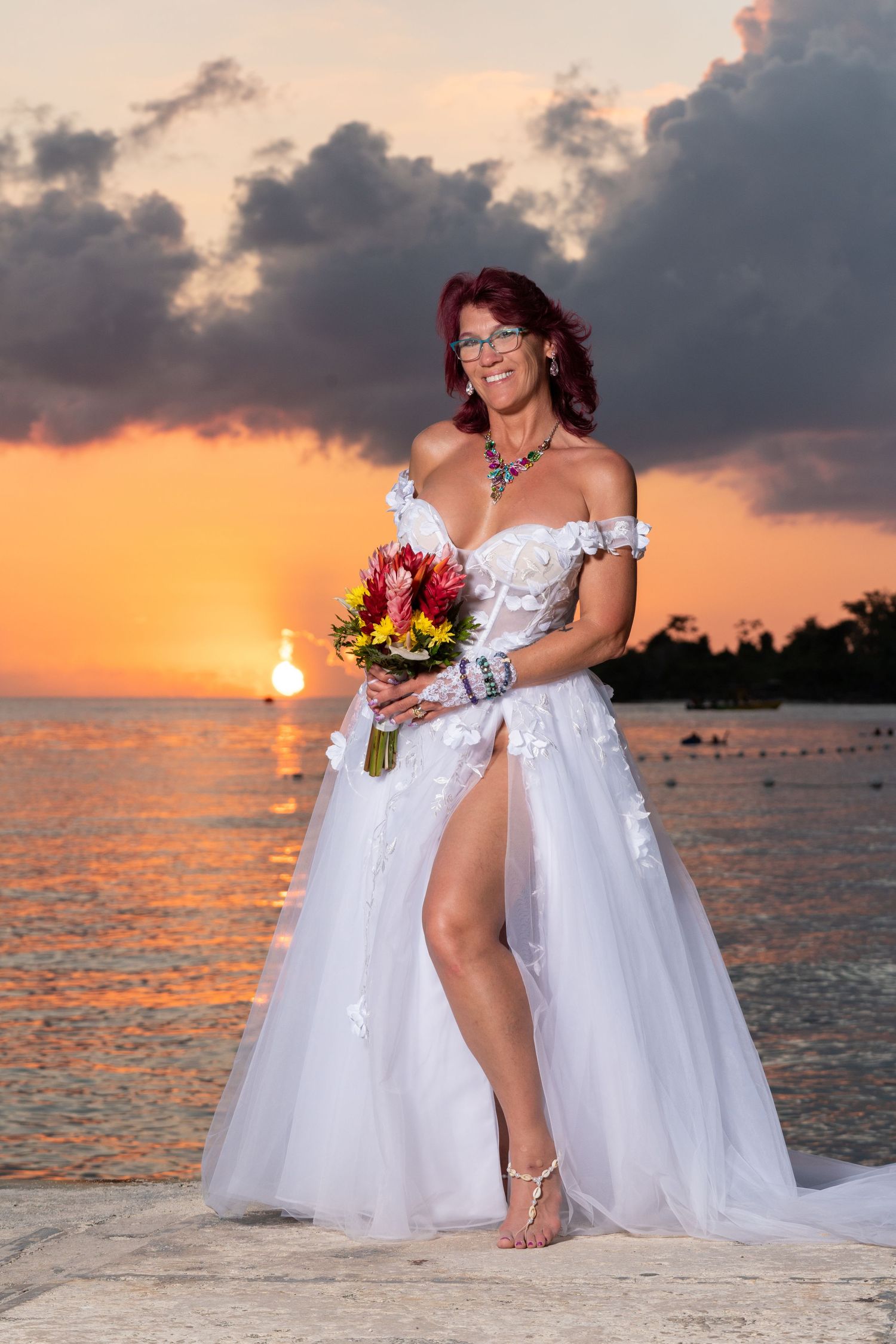 Sunset beach wedding portrait with white off-shoulder dress, colorful bouquet and dramatic orange sky over ocean.