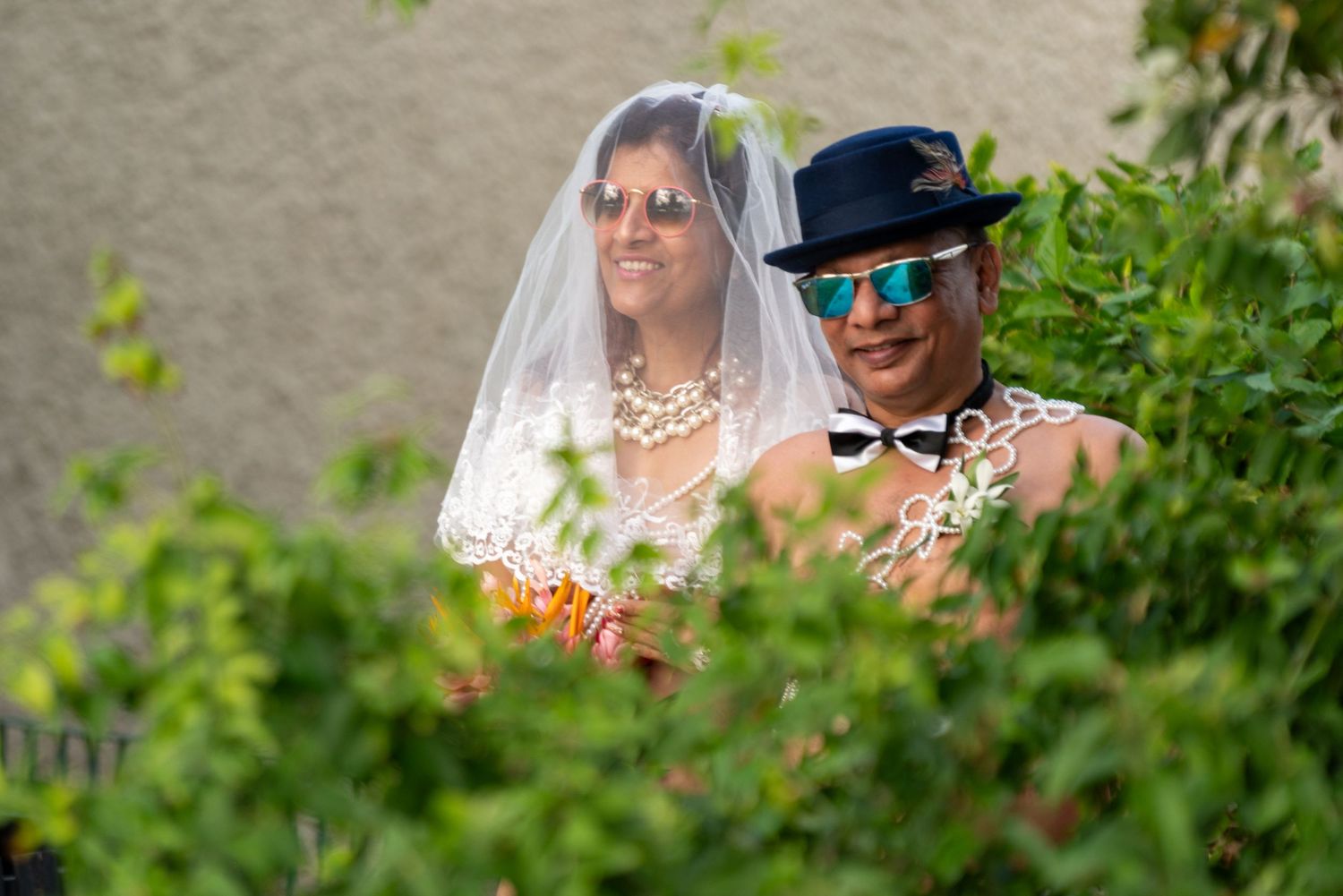 Wedding couple wearing sunglasses peek through green foliage during outdoor celebration.