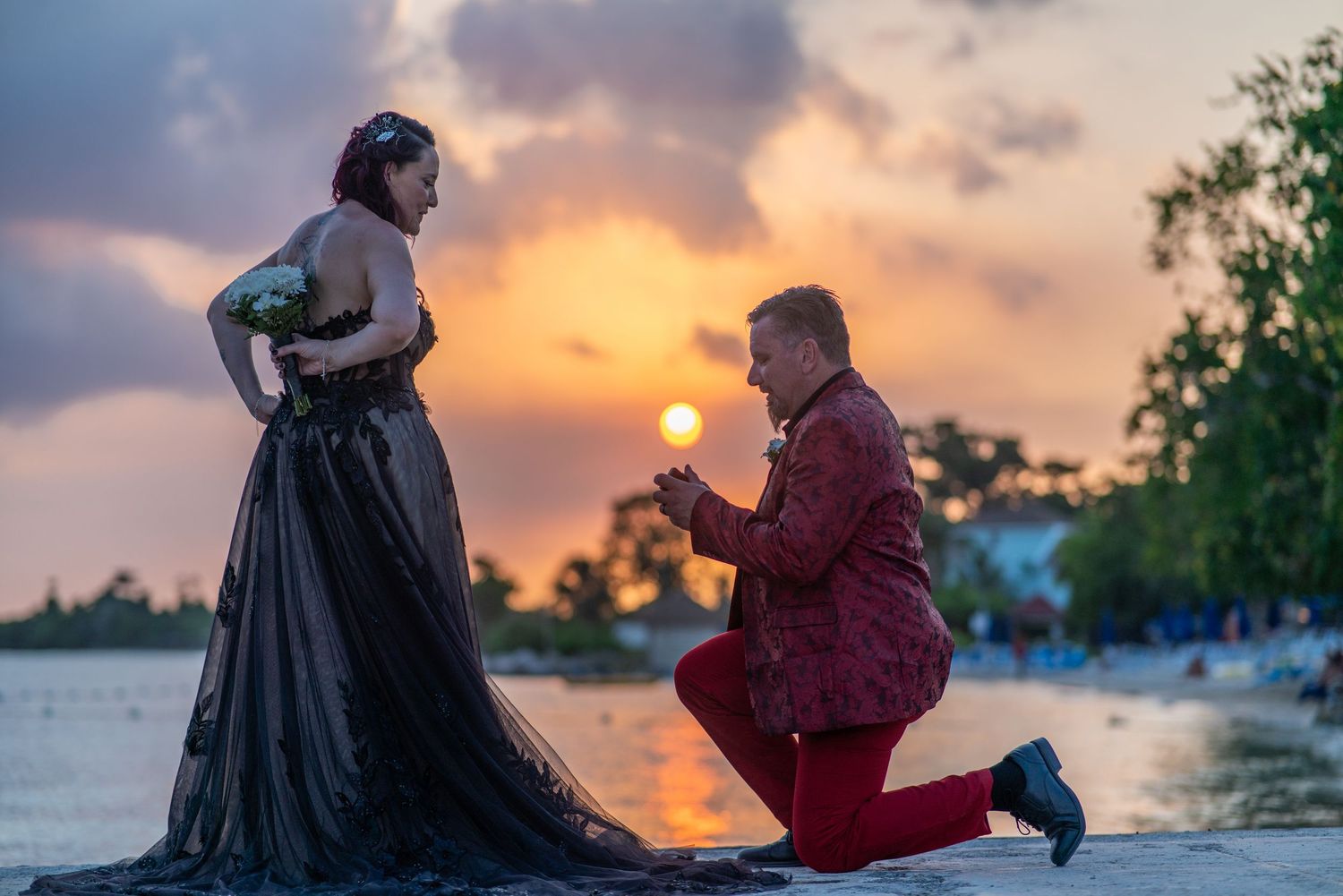 Silhouettes of a romantic sunset beach proposal with soft orange clouds and palm trees in the background.