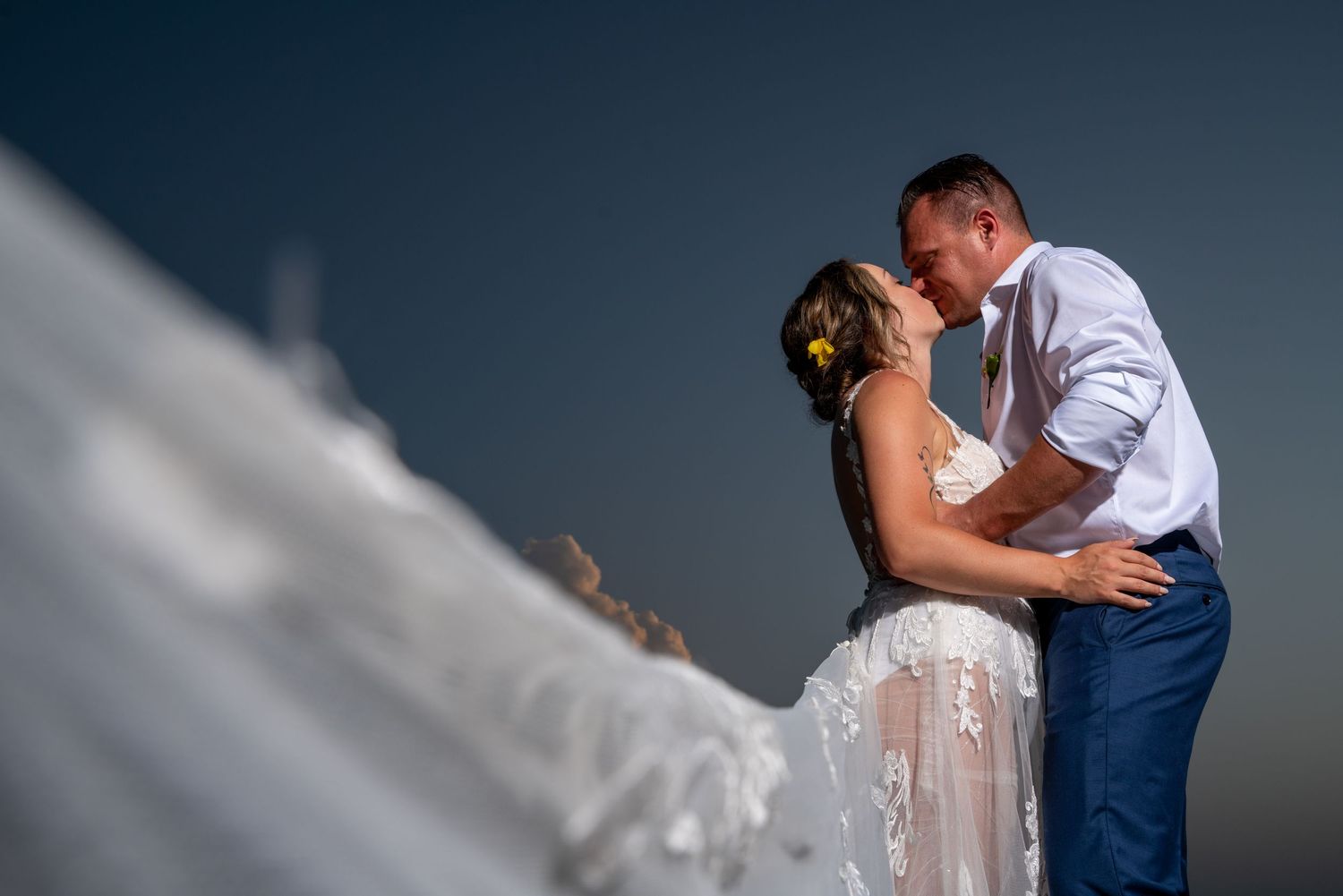 A romantic wedding couple shares a passionate kiss on a scenic overlook against a dramatic blue sky.