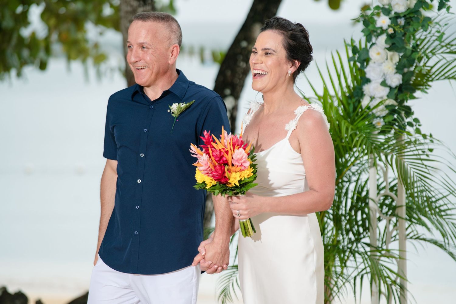 A beach wedding couple stands together holding a vibrant tropical bouquet with pink flowers on a sunny day.