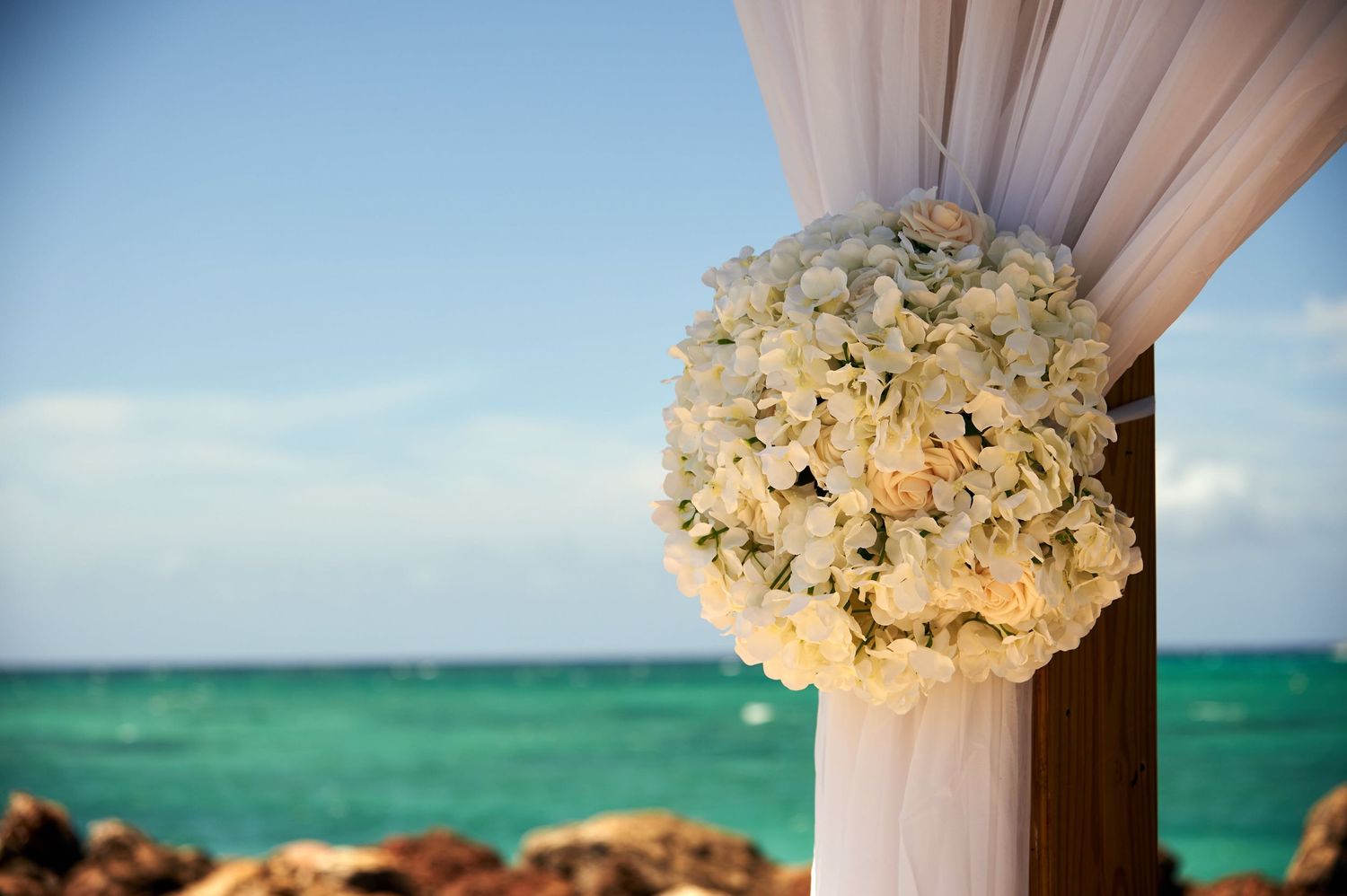 A large white rose bouquet adorns a draped pillar against a turquoise ocean backdrop at a beach wedding ceremony.