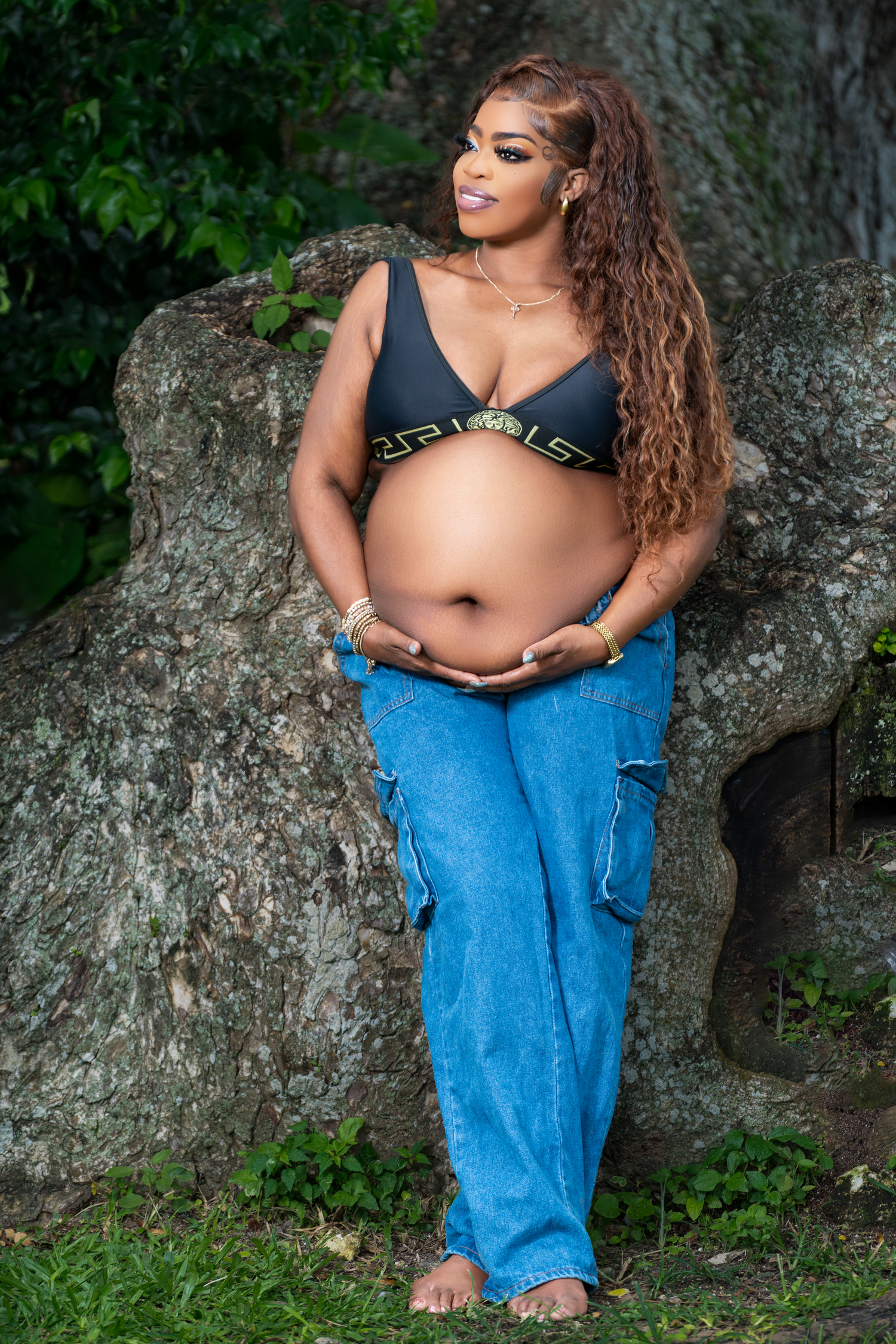 Maternity photoshoot on a rock formation in nature wearing denim and black top against lush greenery.