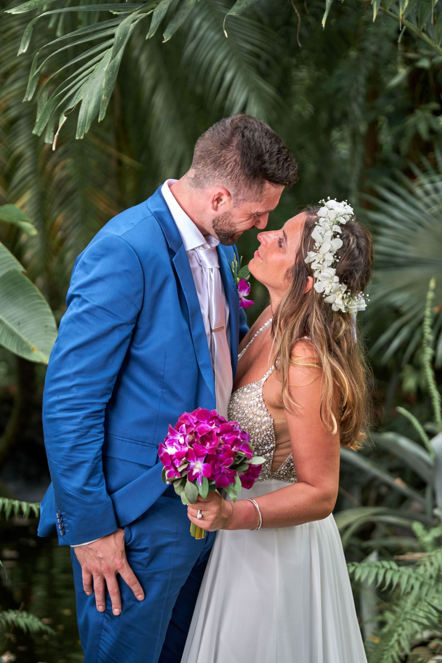 A romantic wedding moment in a tropical garden setting with vibrant pink orchid bouquet and palm fronds in the background.