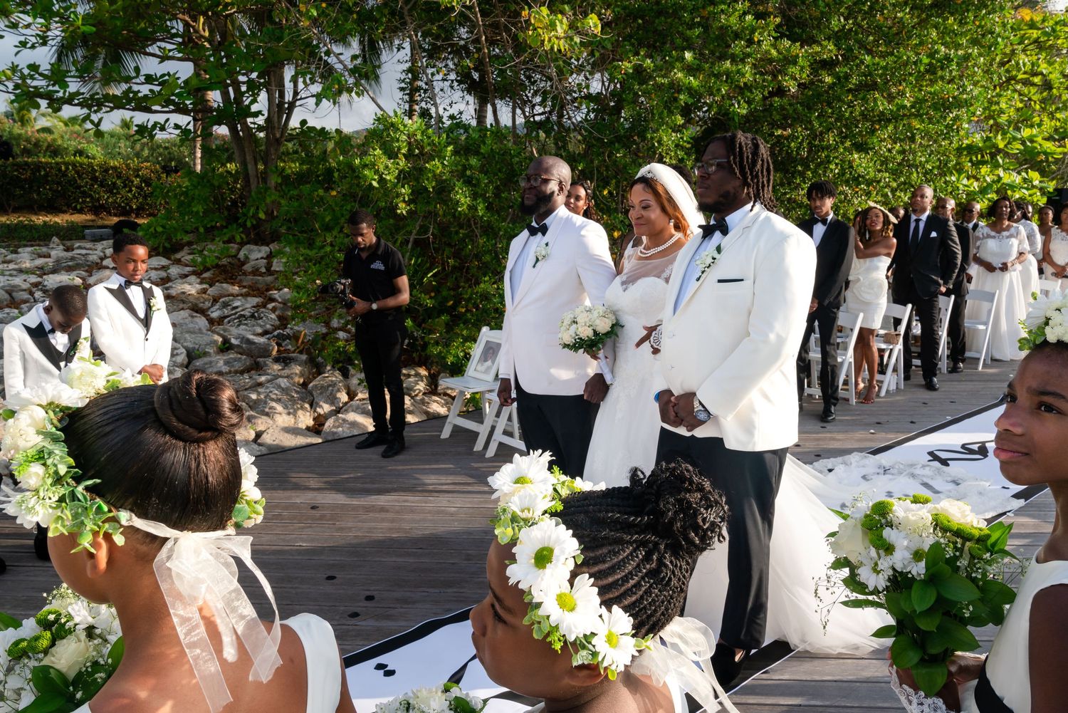 Outdoor wedding ceremony with guests seated in white chairs along a decorated aisle.