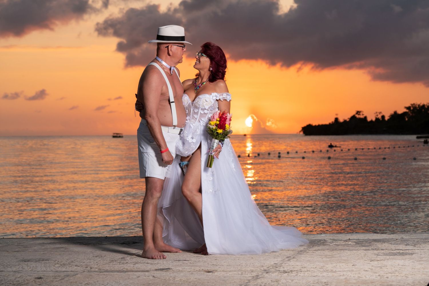 A couple in wedding attire stands barefoot on a beach at sunset with orange sky and calm ocean waters in the background.