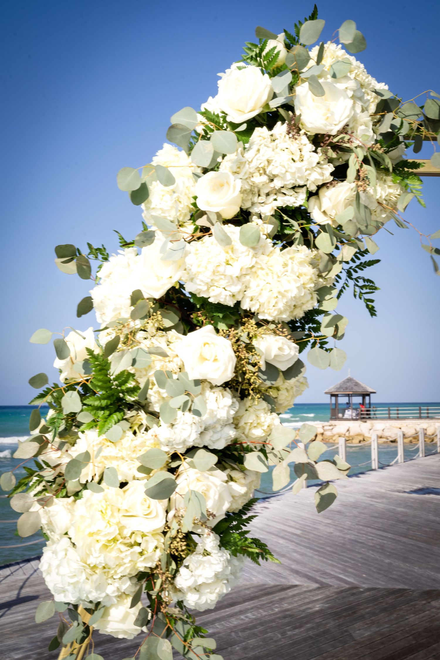 Elegant white floral arrangement with roses and greenery beside a tropical beach gazebo and ocean view.