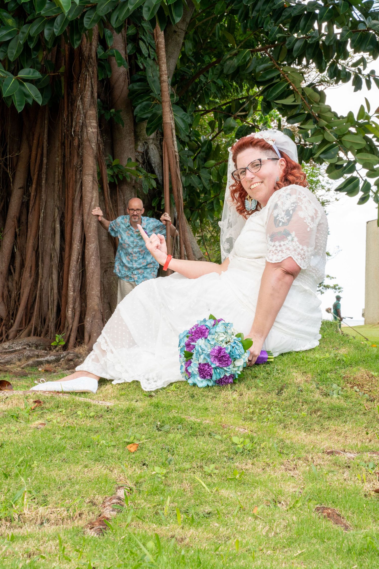 A bride in a white lace dress sits on grass near a large tree trunk while holding a blue and purple bouquet.