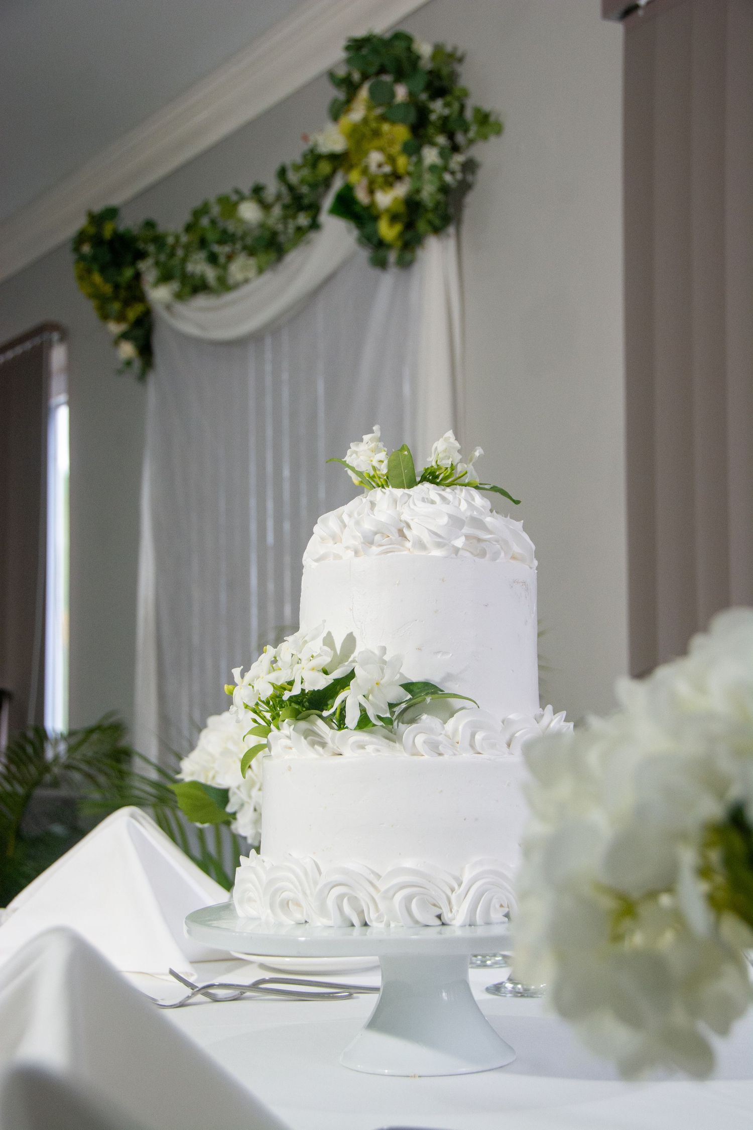 Elegant two-tier white wedding cake with floral decorations and swirled frosting details displayed on pedestal stand.