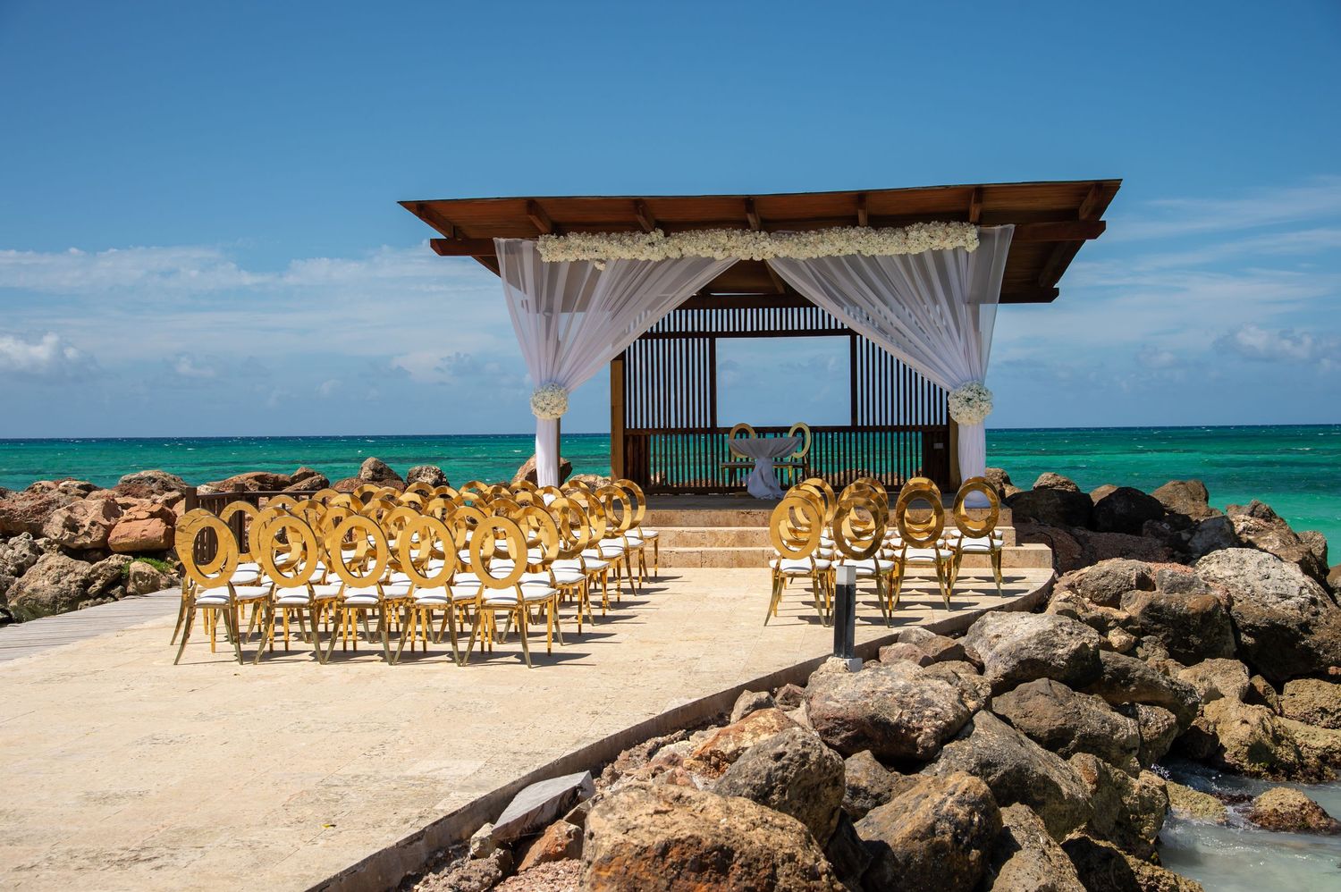 Rows of wooden chairs set up for a beach wedding ceremony with turquoise ocean and rocky coastline views.