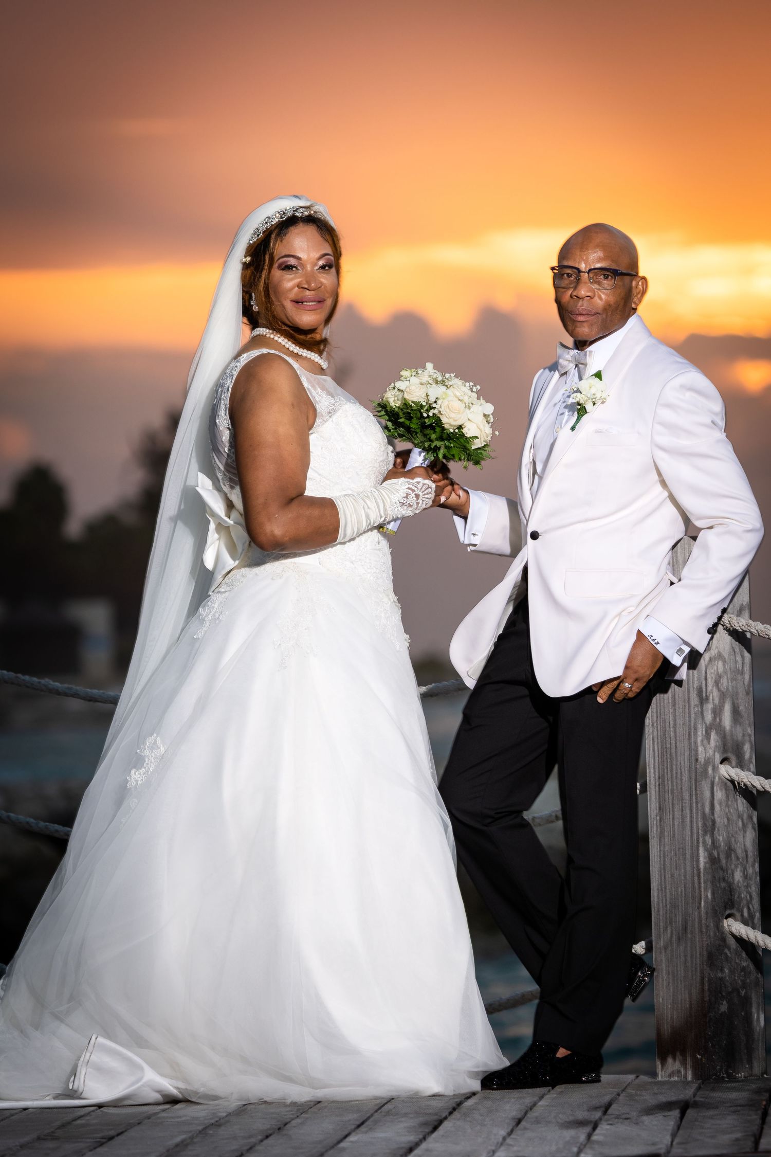 Newlywed couple poses together against vibrant orange sunset sky at outdoor waterfront location.