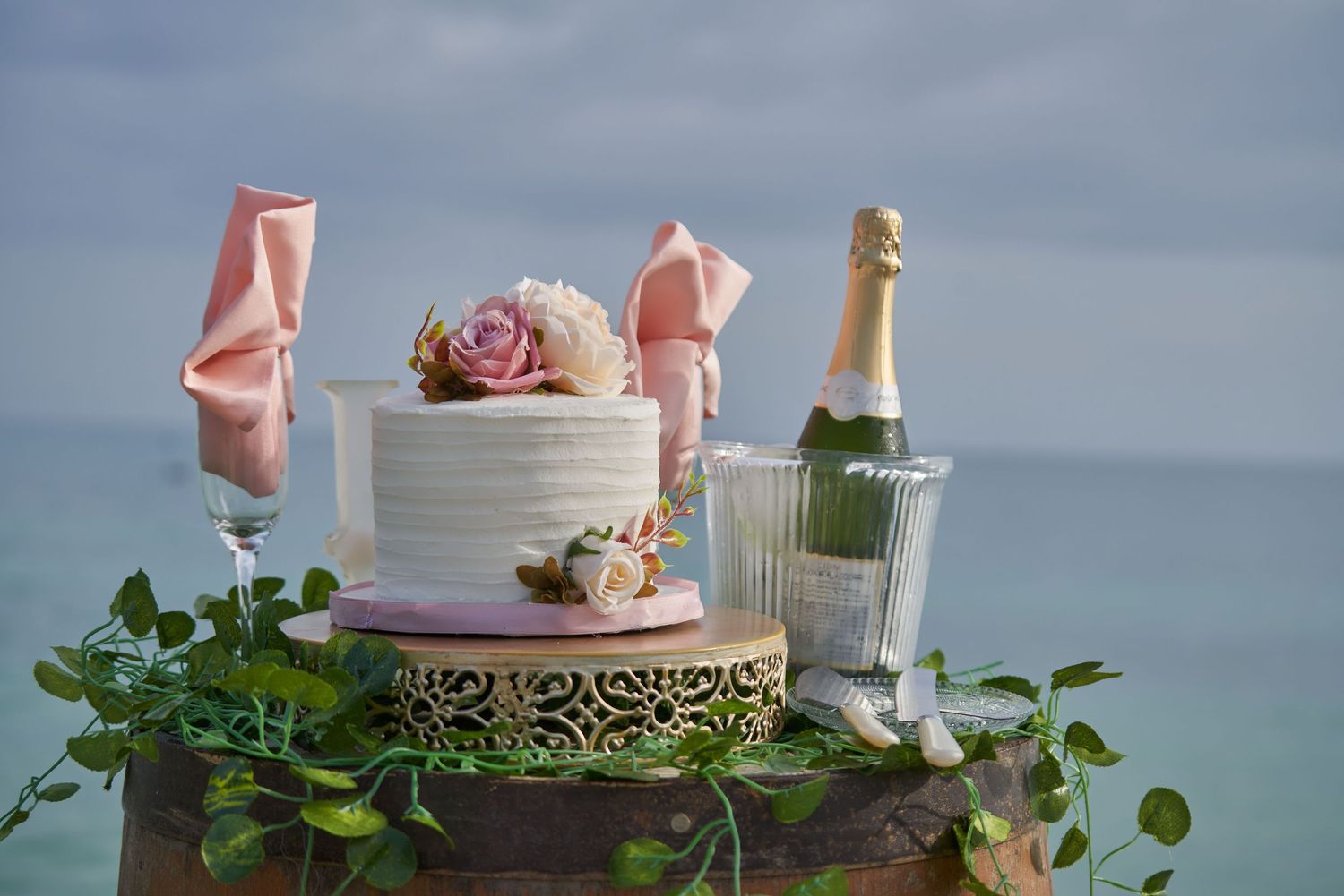 Elegant pink and gold wedding cake decorated with flowers and roses sits on a tiered stand against a blue sky background.