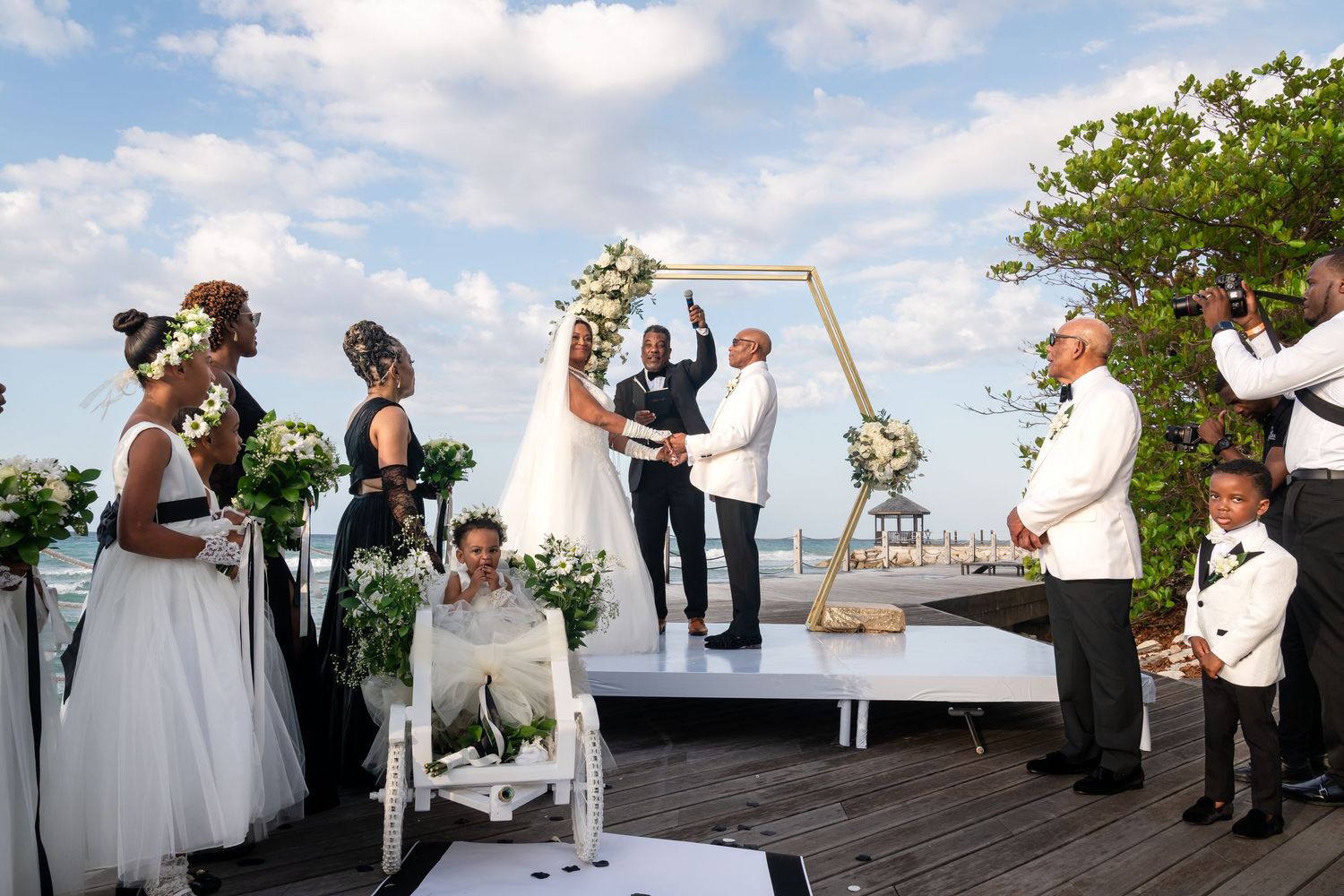 A wedding ceremony takes place at a waterfront venue with a wooden arch setup against the ocean backdrop.