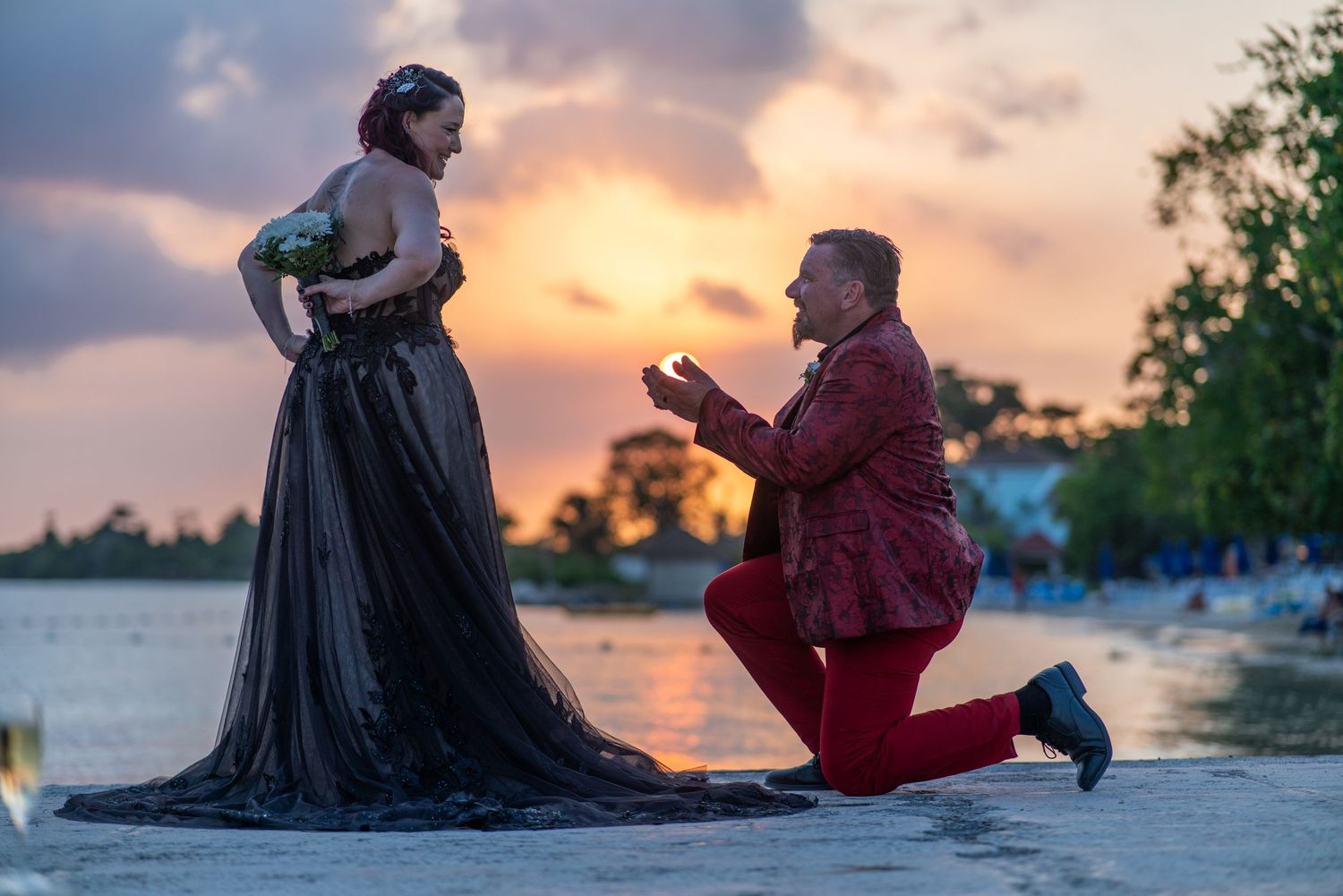 A couple shares a romantic moment during a beach proposal at sunset with dramatic orange sky in the background.