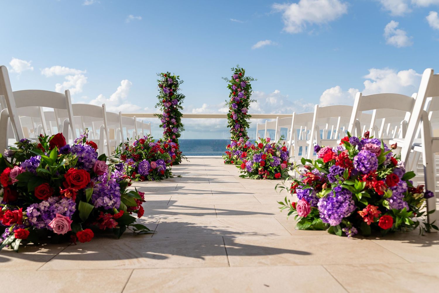 White chairs line a flower-decorated aisle leading to an ocean view wedding ceremony setup on a sunny day.