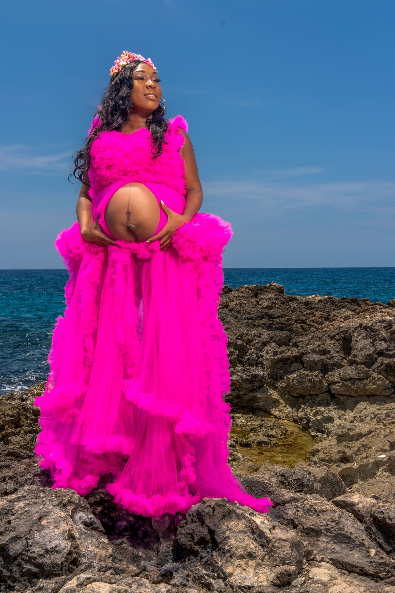 Expecting mother in a vibrant hot pink tulle dress cradling her belly against rocky coastal cliffs and blue ocean backdrop.