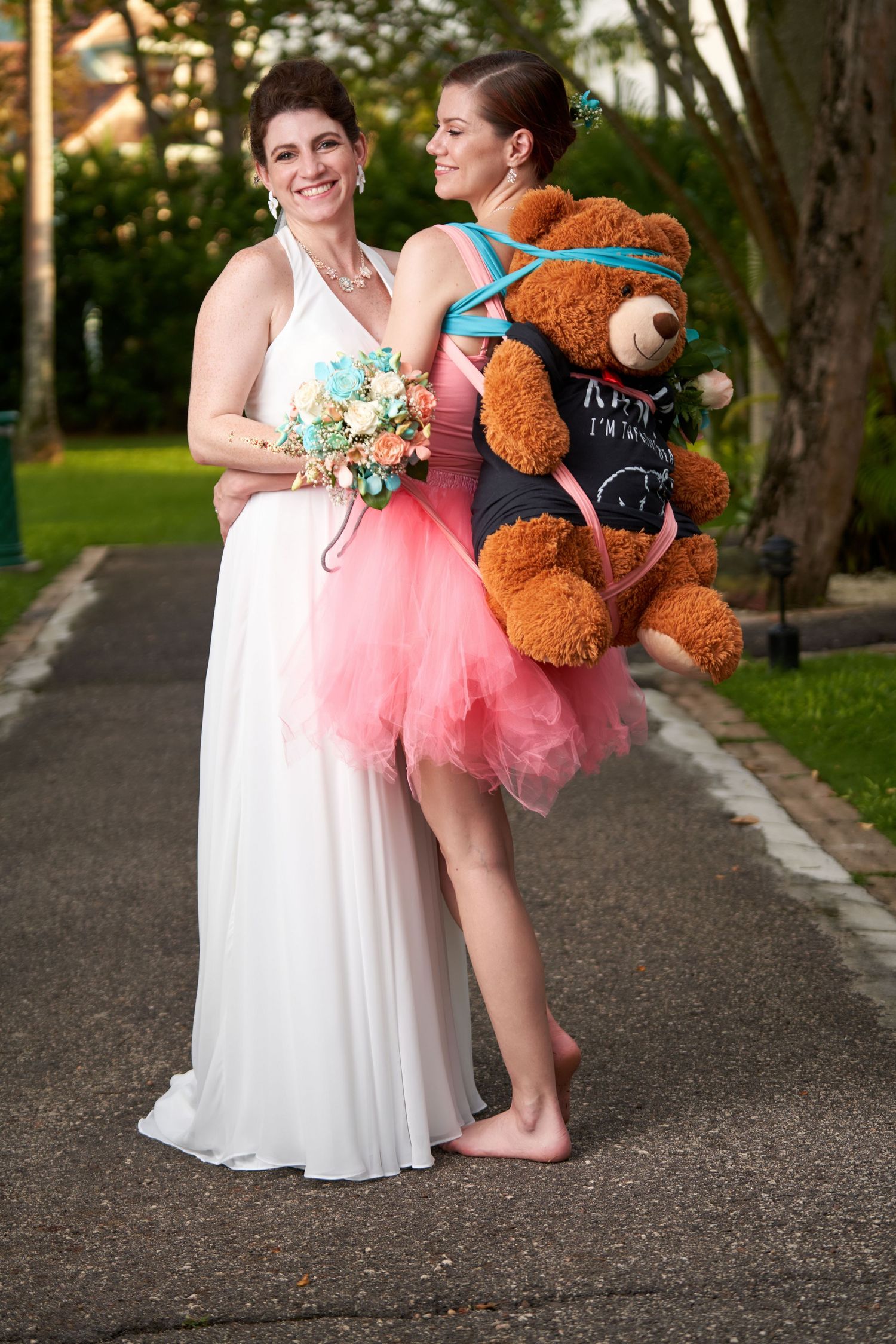 Bride in white wedding dress shares a tender moment with bridesmaid who has on a pink dress and a teddy bear backpack.