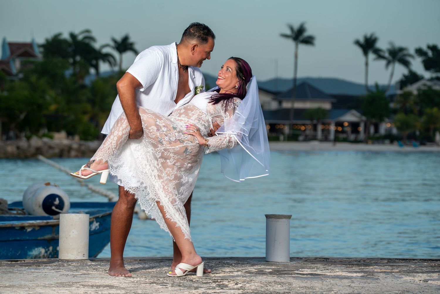 A couple in pink wedding attire leans romantically together on a dock overlooking tropical blue waters.