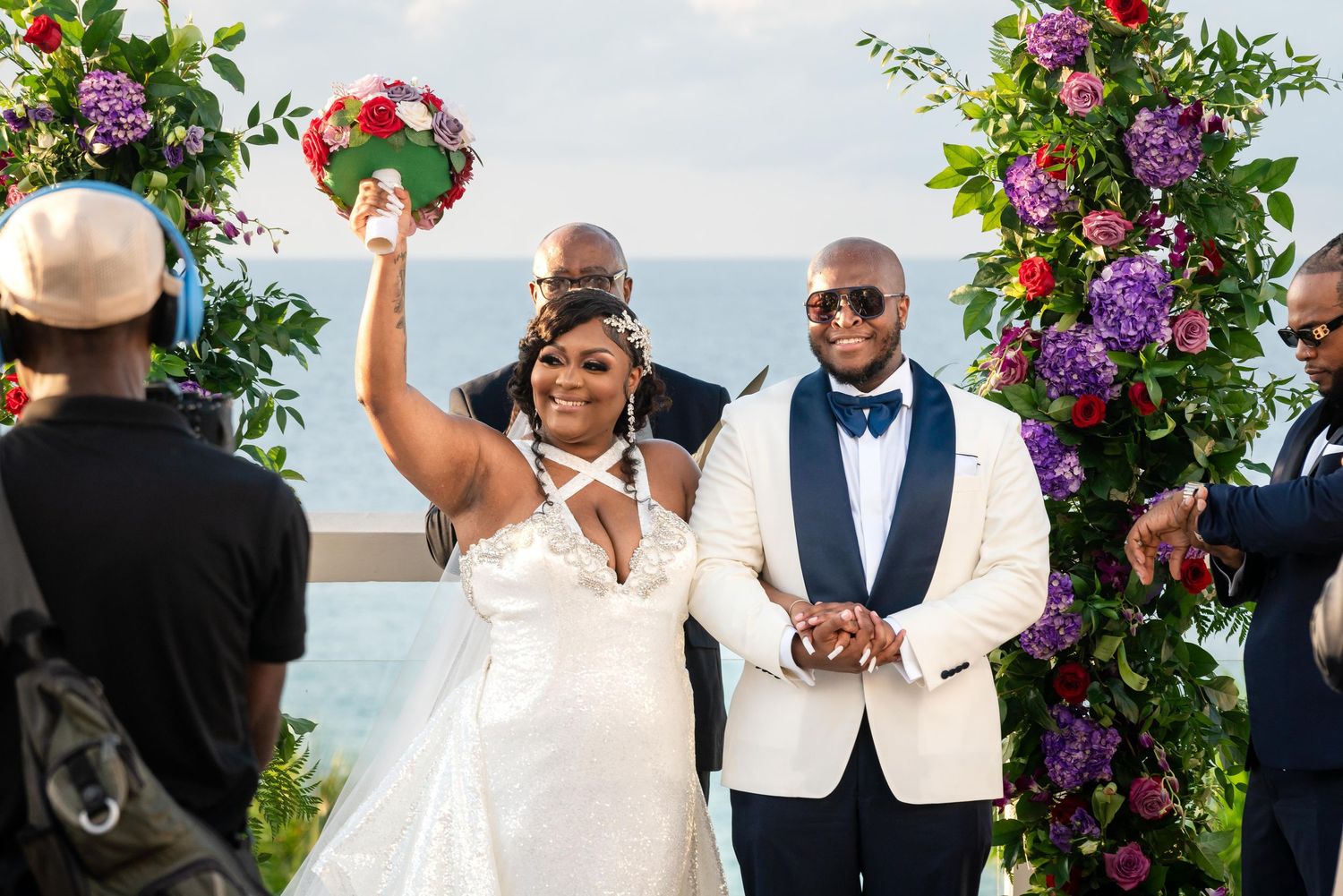 Elegant wedding celebration on a beach with vibrant purple flowers and greenery decorating the outdoor ceremony space.