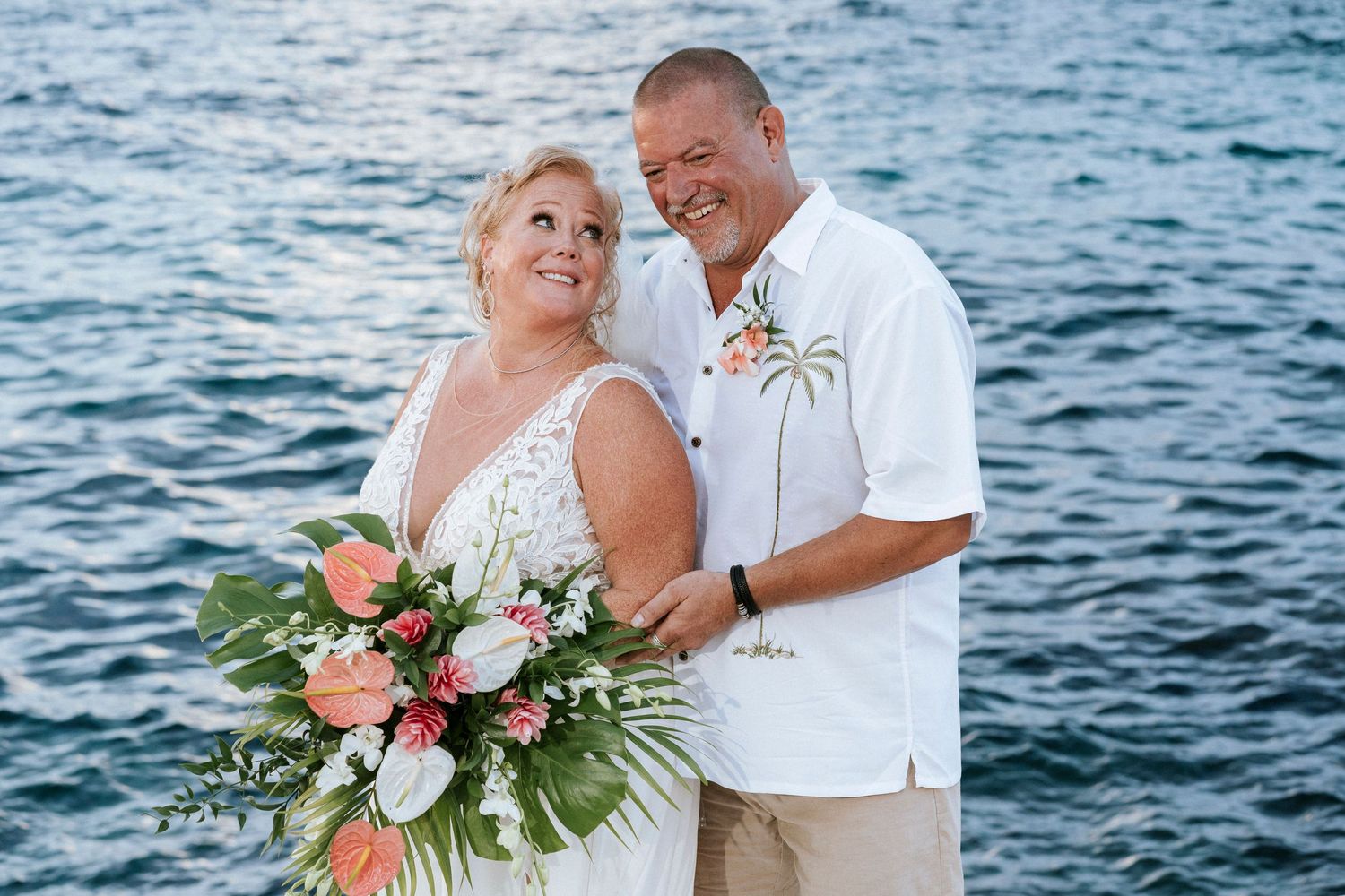 Happy couple embraces on beach with coral floral bouquet and blue ocean waters in background.