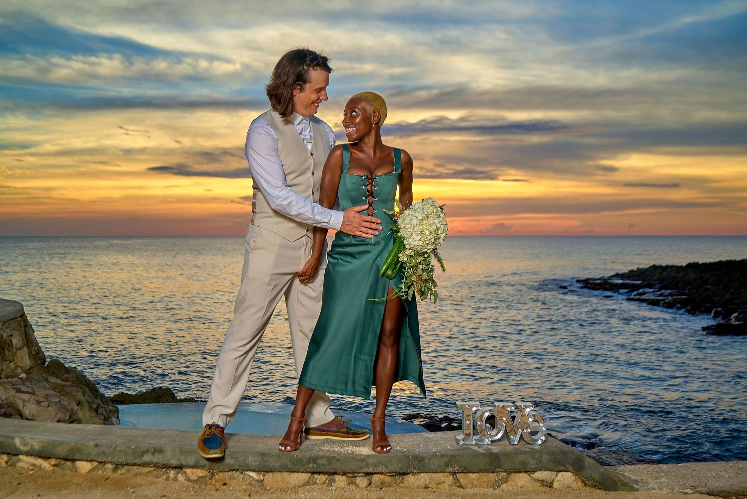 Couple in wedding attire embracing at sunset on rocky Hawaii coastline with vibrant orange sky and ocean backdrop.