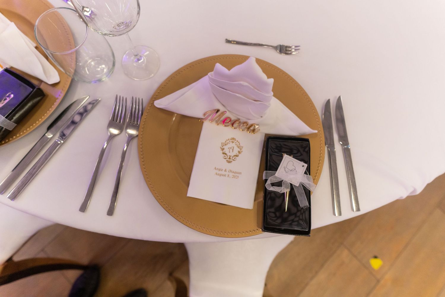 Place settings of gold charger plates, silverware, and wedding menu cards arranged on a white tablecloth for a formal event.