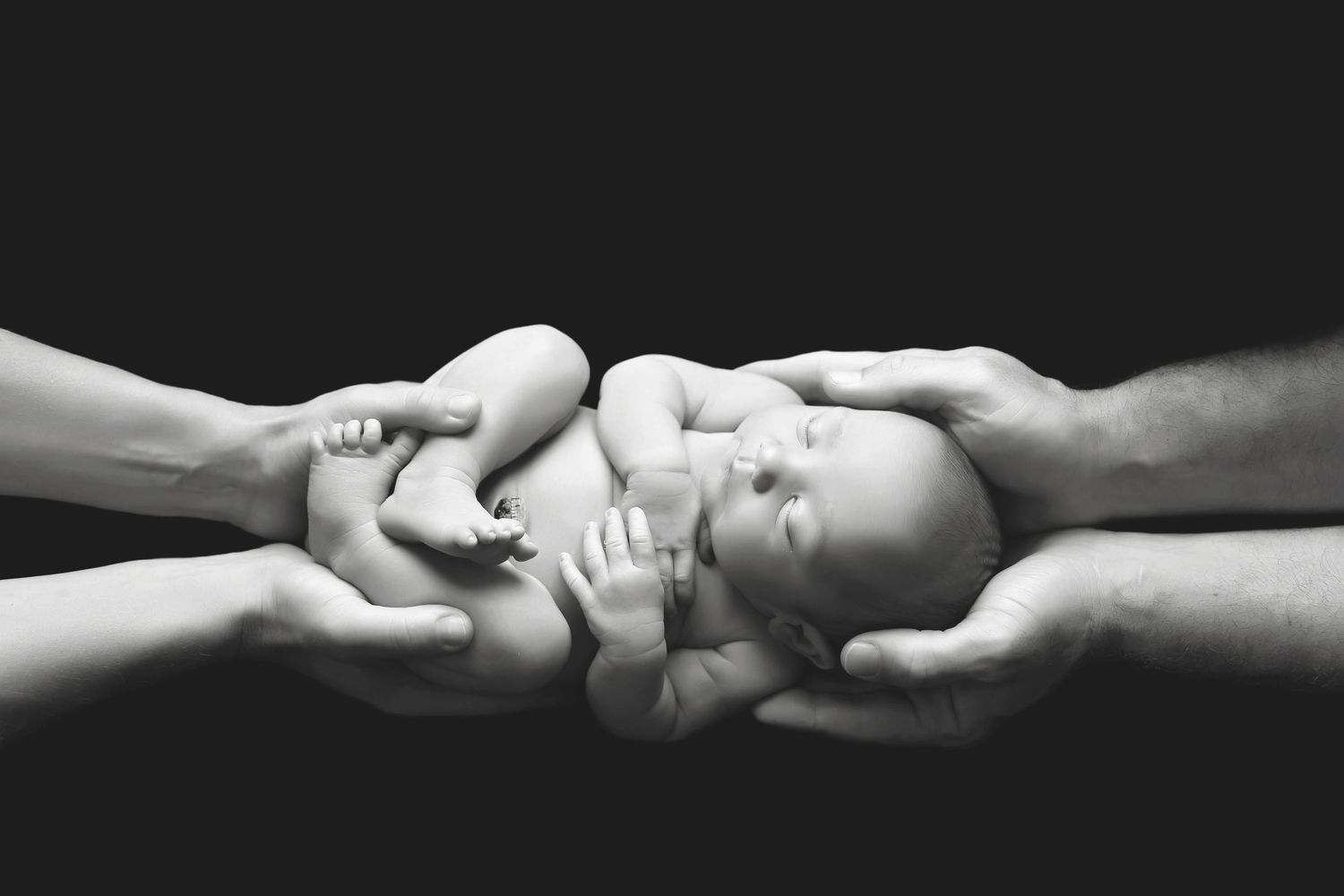 Black and white photo of a newborn baby cradled gently in the hands of two adults against a dark background.