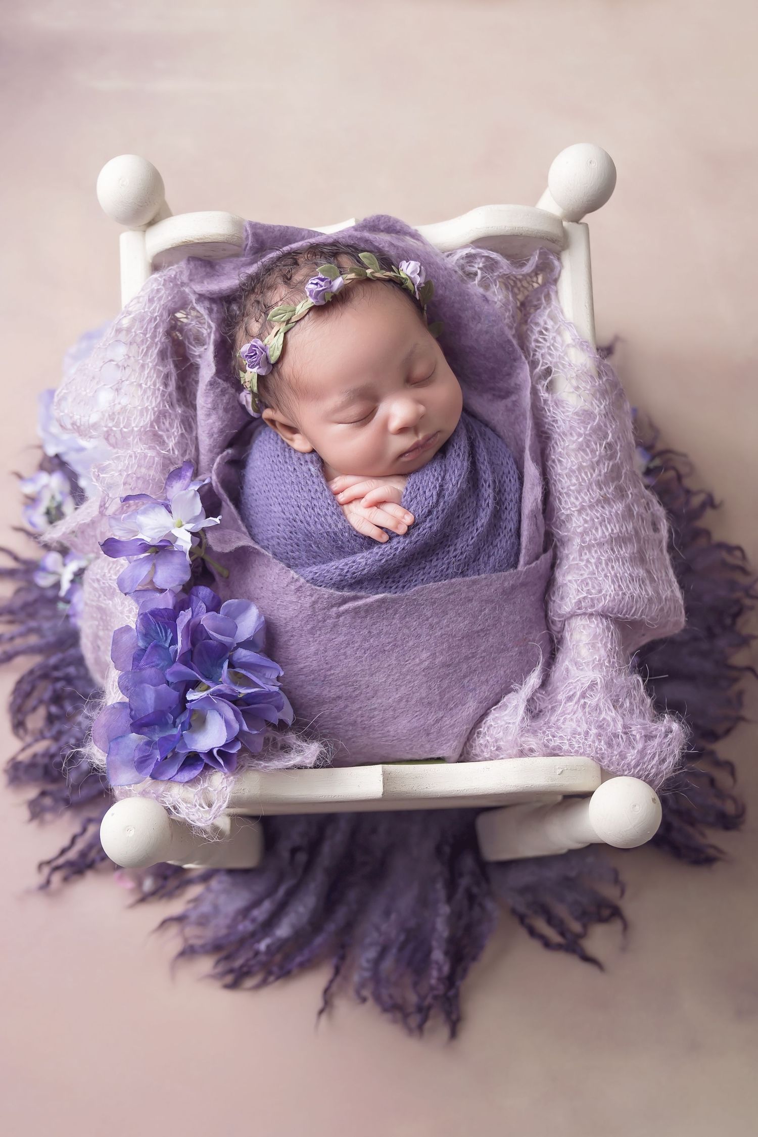 Baby sleeping on miniature white bench surrounded by purple flowers and textured fabric. San Diego Newborn Photographer