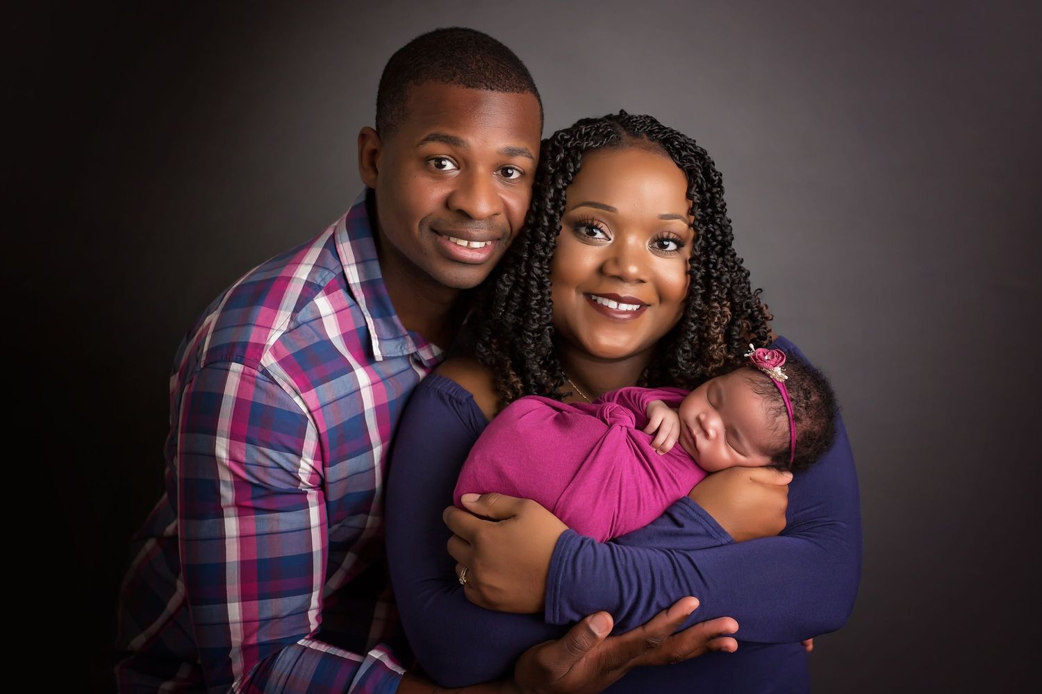 Studio portrait of family with baby in pink wrap against dark background. San Diego Newborn Photographer