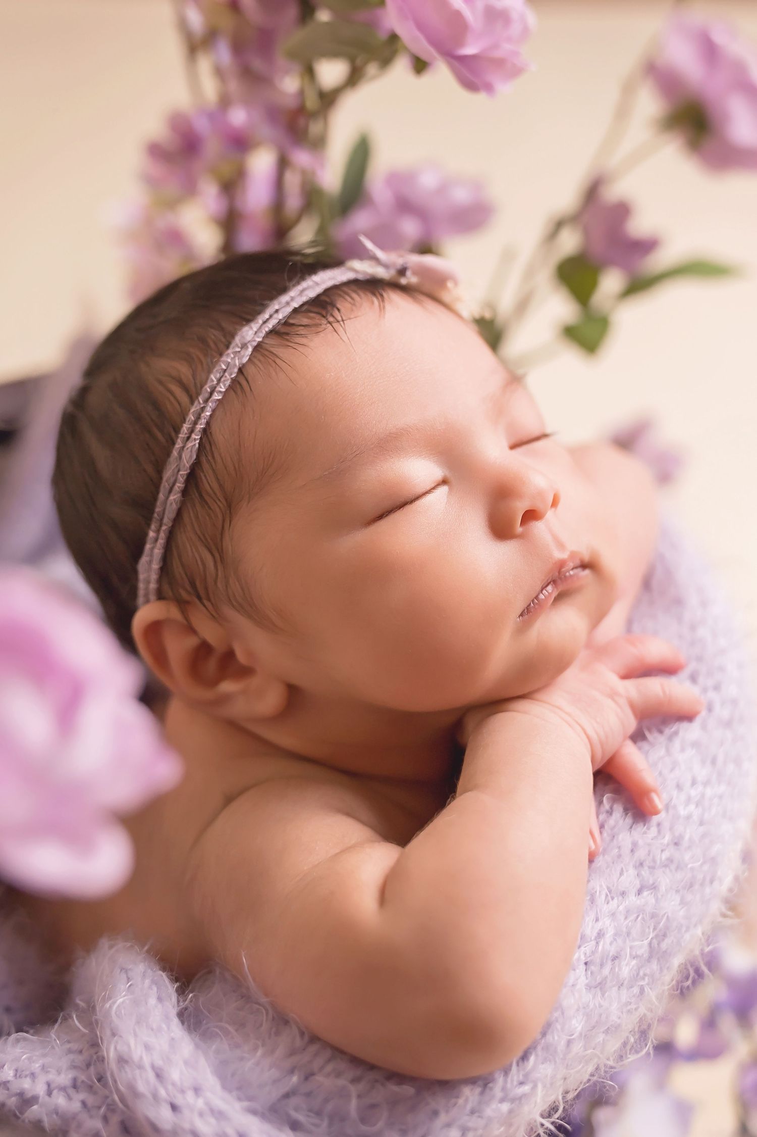 Close-up of sleeping newborn with purple flowers and lace wrap against neutral backdrop. San Diego Newborn Photographer