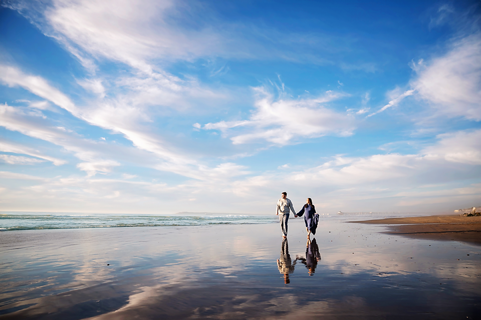 Couple holding hands walking on a reflective wet beach with dramatic blue sky and wispy clouds at sunset.