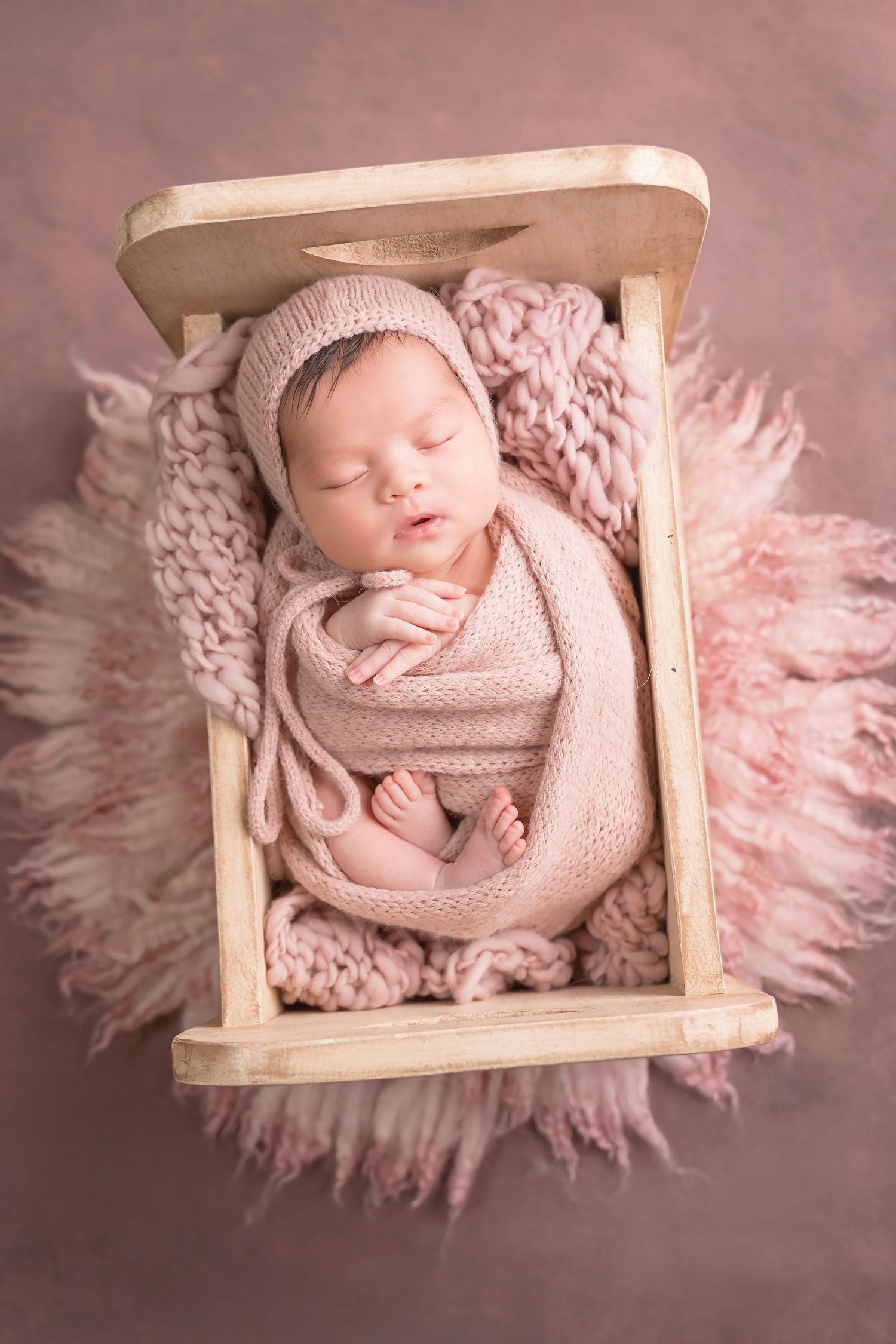 Sleeping newborn wrapped in pink knit blanket nestled in wooden crate with fluffy pink accents. San Diego Newborn Photographer