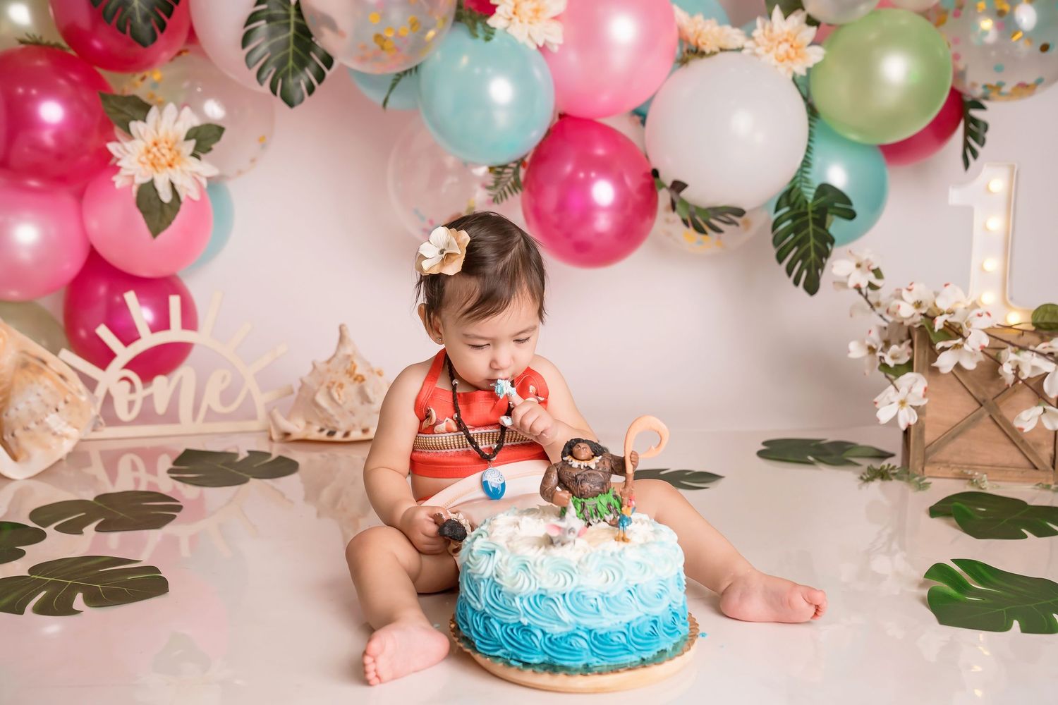 Festive birthday scene with colorful balloons, tropical leaves and a blue frosted cake.