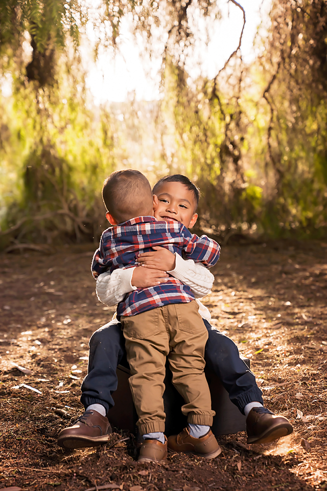 Two young brothers share a warm hug outdoors, smiling joyfully in a sunlit park setting.