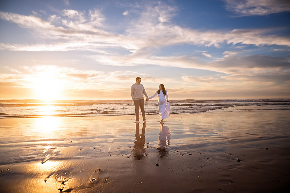 Couple holding hands on a reflective beach at golden sunset with dramatic sky and ocean waves.