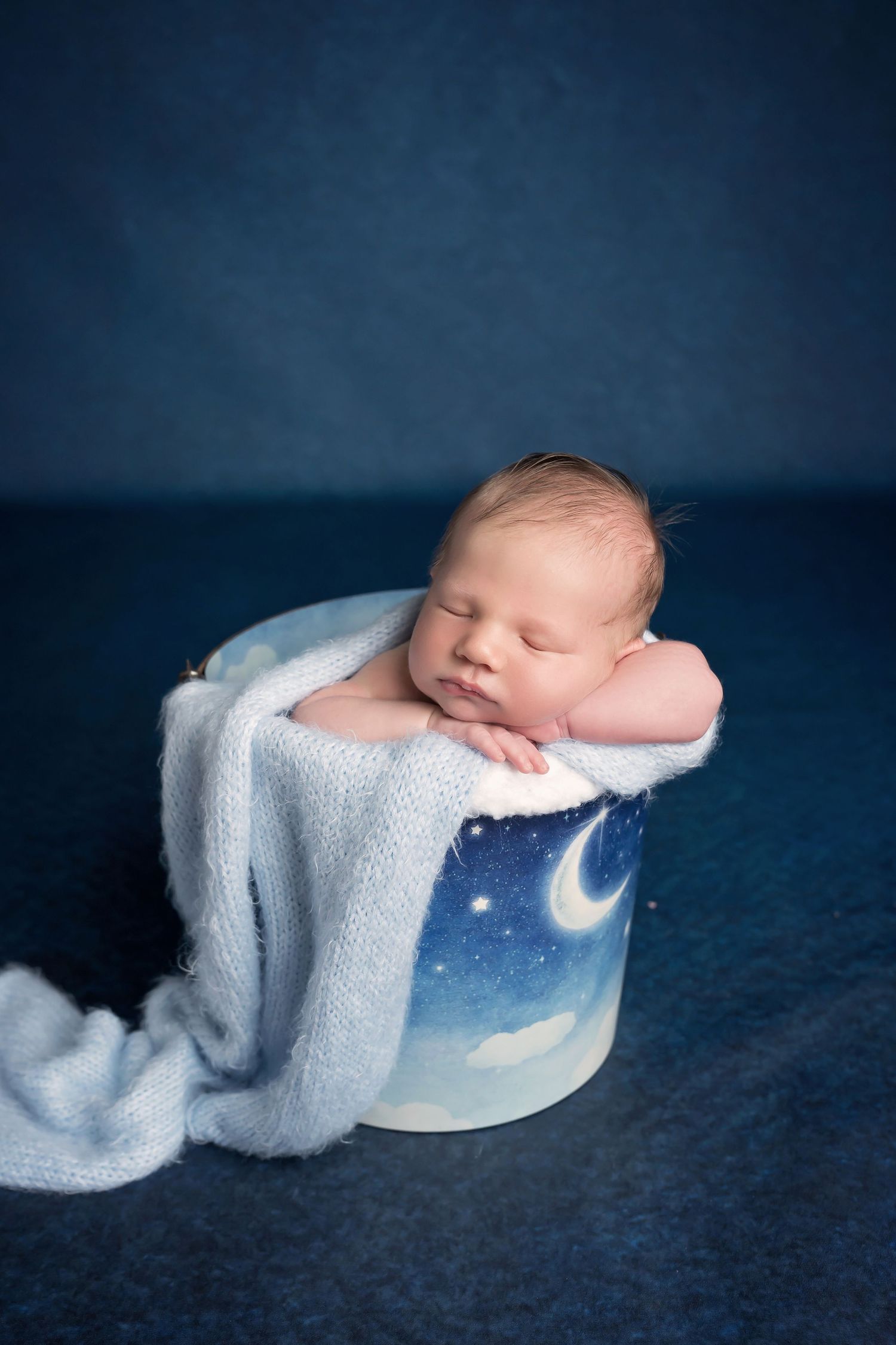 Sleeping newborn on moon-themed round prop with soft blue background and white wrap. San Diego Newborn Photographer