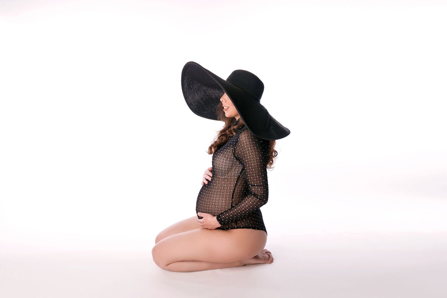 Artistic maternity pose with person wearing black hat and sheer top in minimalist setting.