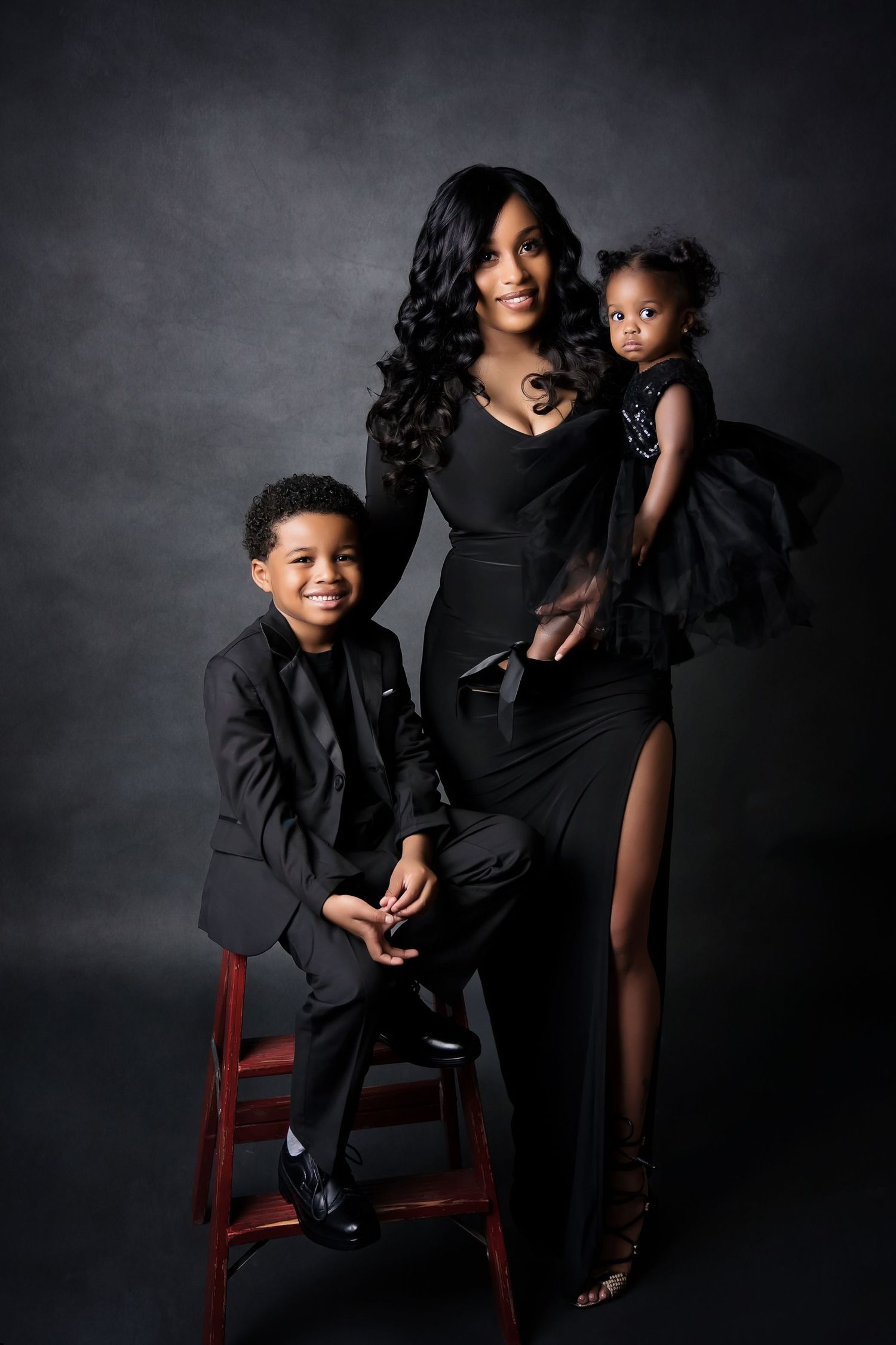 Studio portrait of family in elegant black outfits posed with a red chair against a dramatic dark backdrop