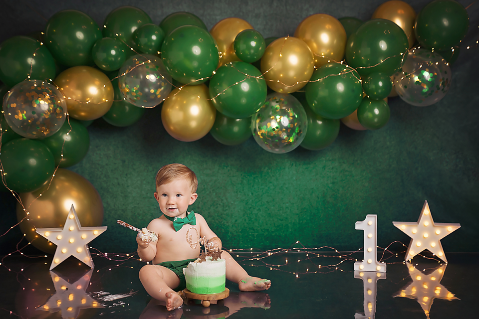 Baby's first birthday cake smash with green and gold balloons, star decorations, and number one marquee light on dark background.