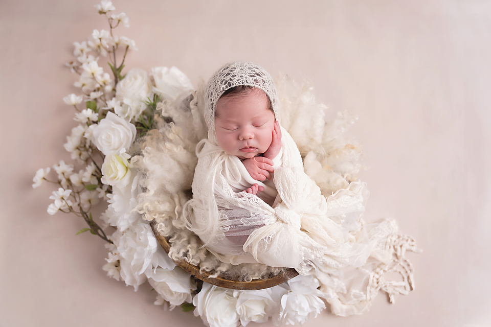 Sleeping newborn baby wrapped in cream lace swaddle, resting in a bowl with white flowers on a pink background.