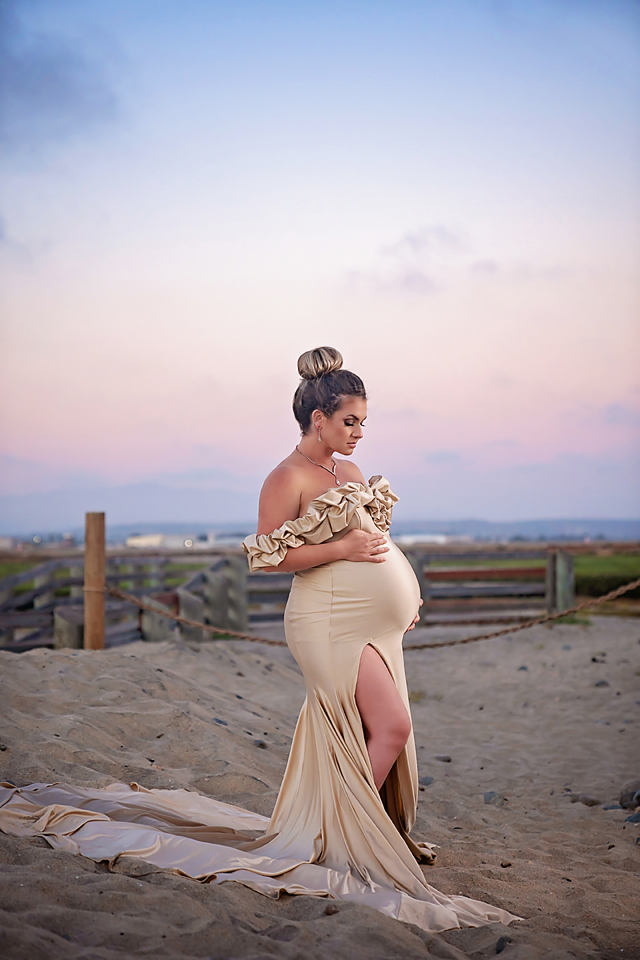 Pregnant woman in elegant gold off-shoulder gown poses on sandy beach at sunset during maternity photoshoot.