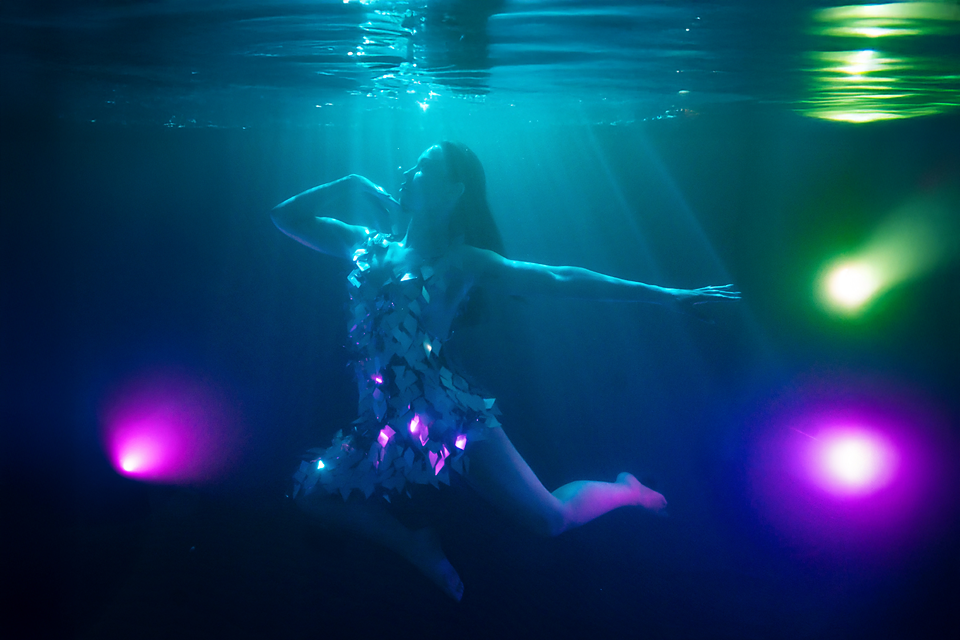 Woman in glowing dress floating underwater surrounded by colorful pink and green lights in dark blue water.
