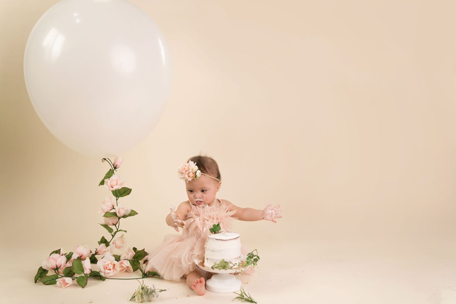 Baby in pink dress poses with floral decorations and white balloons against cream backdrop. San Diego cake smash photographer