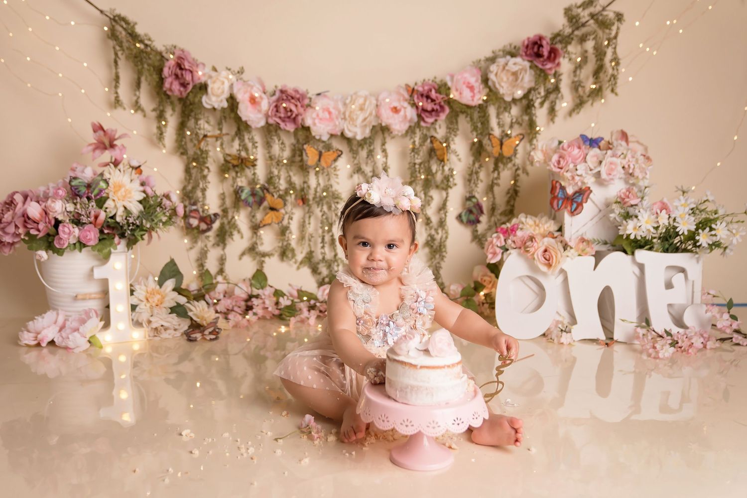 Pink floral first birthday setup with cake and white letter decorations on beige background.