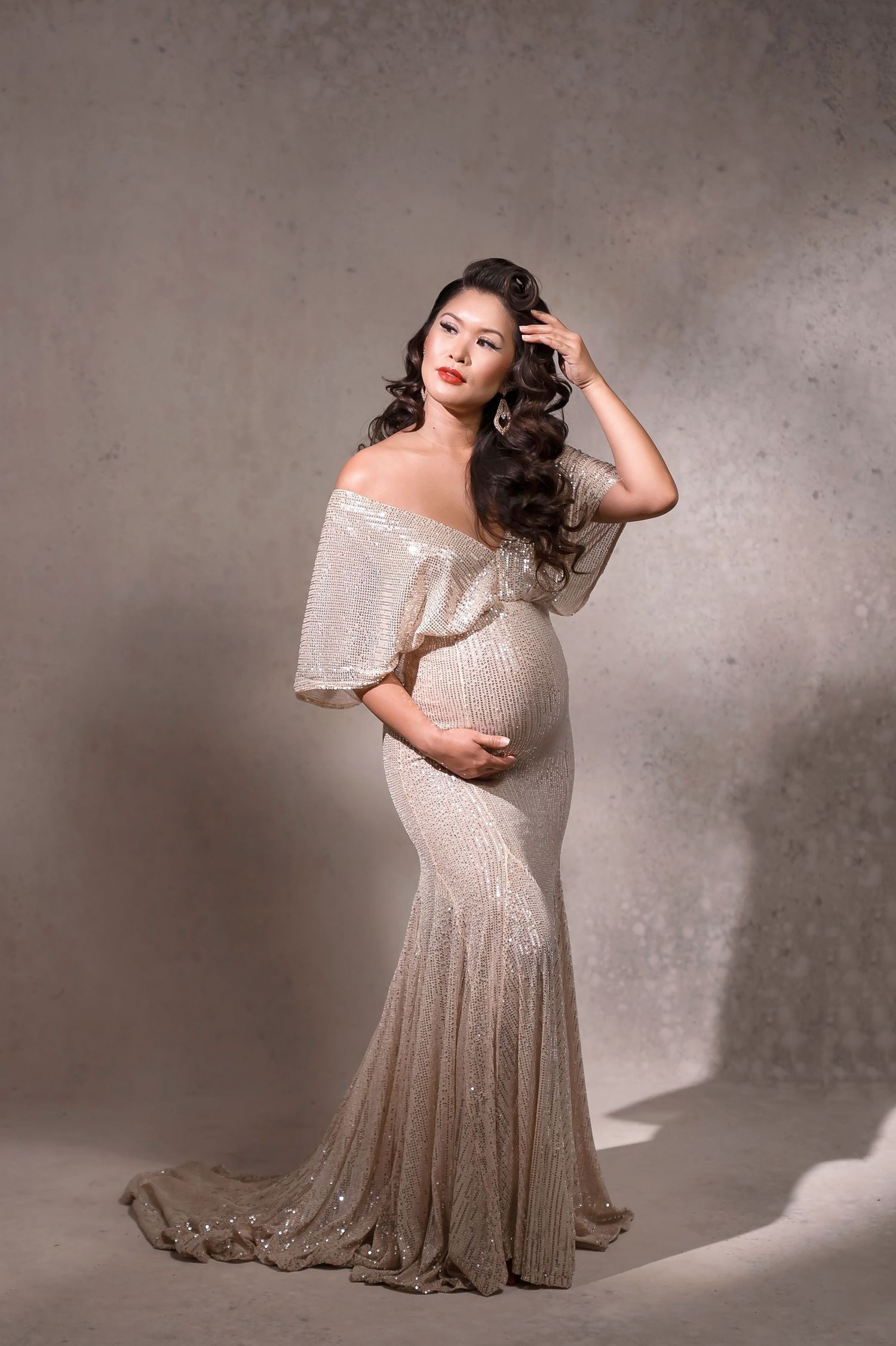 Pregnant woman in elegant off-shoulder sequin gown poses gracefully in studio, showcasing maternity fashion.
