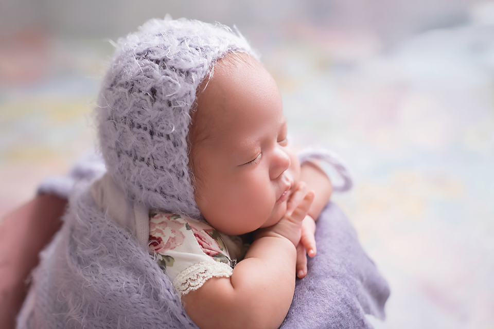 Sleeping newborn baby wearing a soft lavender knit bonnet, wrapped in a purple blanket in a peaceful pose.